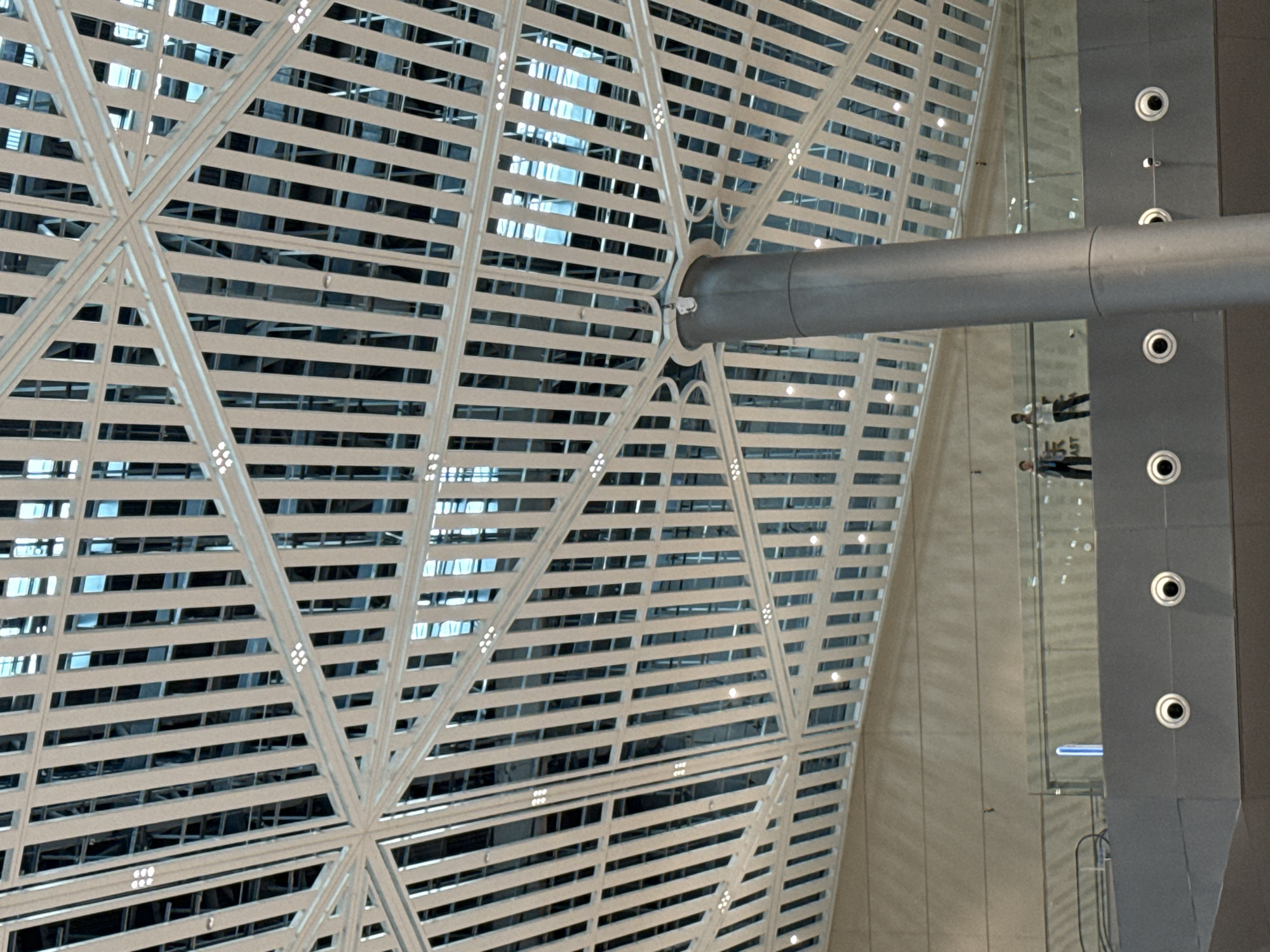 An interior view looking up at a geometric ceiling structure with an attached column in the foreground. The ceiling is comprised of white, angled beams forming a lattice pattern with square openings, revealing a darker interior structure and blue lighting. Two people are visible on a balcony-like structure below the ceiling. One person is wearing a dark shirt and pants, the other a white shirt and dark pants. A blue light fixture is positioned on the balcony railing. The architecture suggests a modern public space, possibly within the Chongqing High-tech Zone, China.