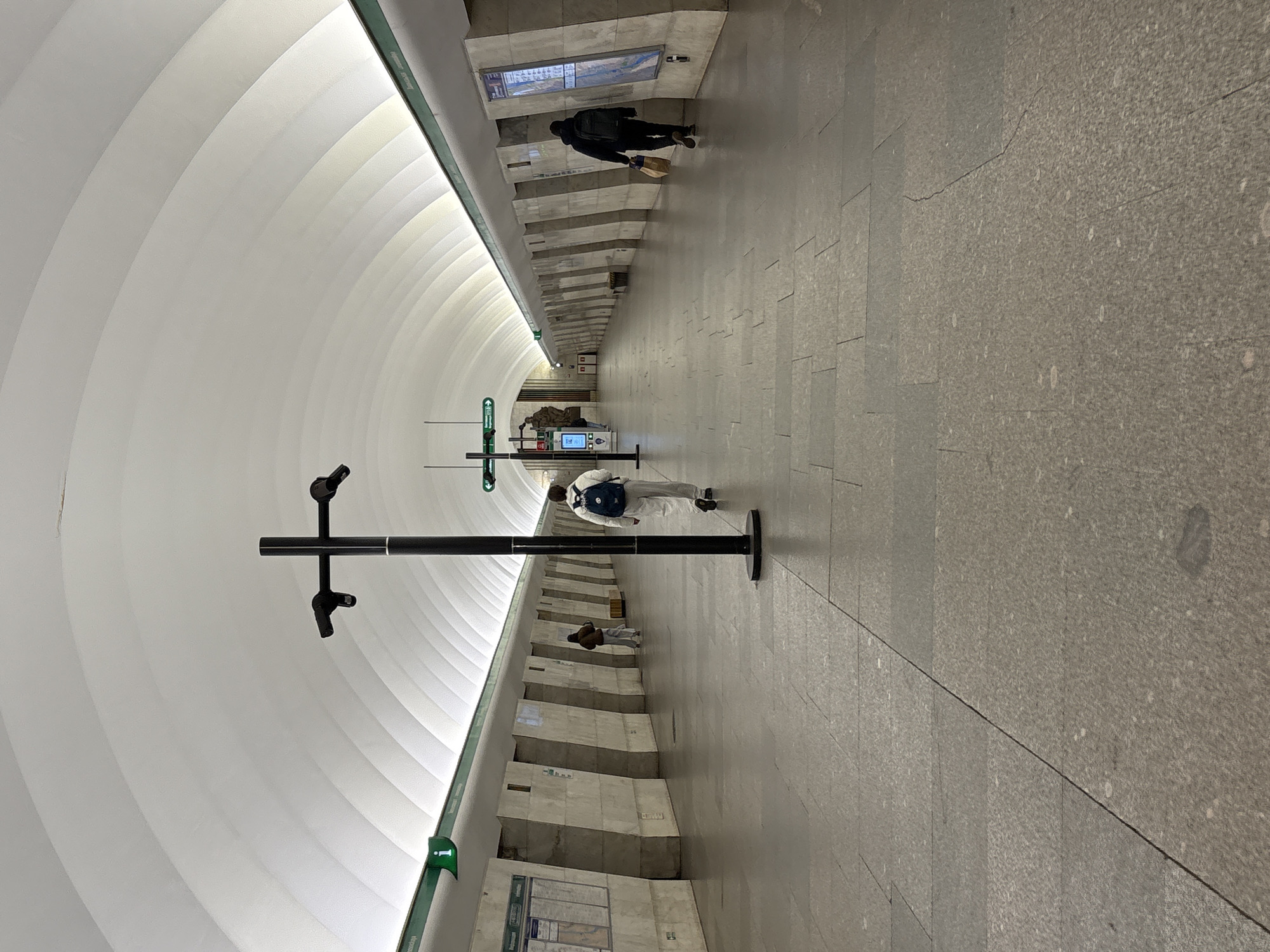 The image depicts a subway station platform in Saint Petersburg, Russia. The station is characterized by its arching, segmented white ceiling. The walls are clad in light gray granite slabs. On the left side, there is a dark gray information pillar, and a few people are seen walking along the platform.