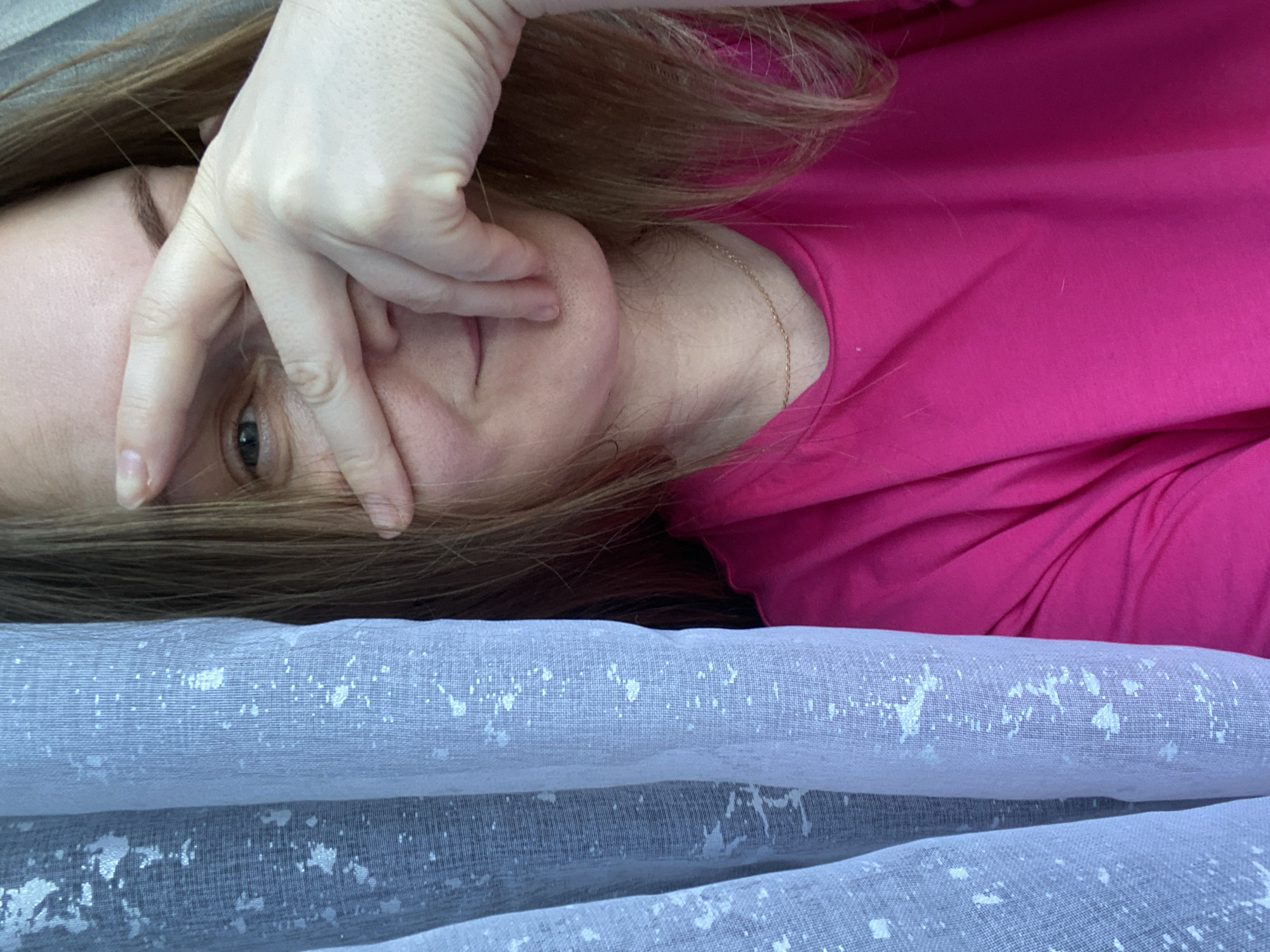 A woman lies on a surface covered with a light blue and white patterned fabric. She wears a bright pink shirt and a delicate gold necklace is visible. She covers part of her face with her hand, revealing only one eye. Her hair is light brown. The background is blurry and provides no context for the location. The image does not contain sufficient details to confirm the location as Noviy, Russia.