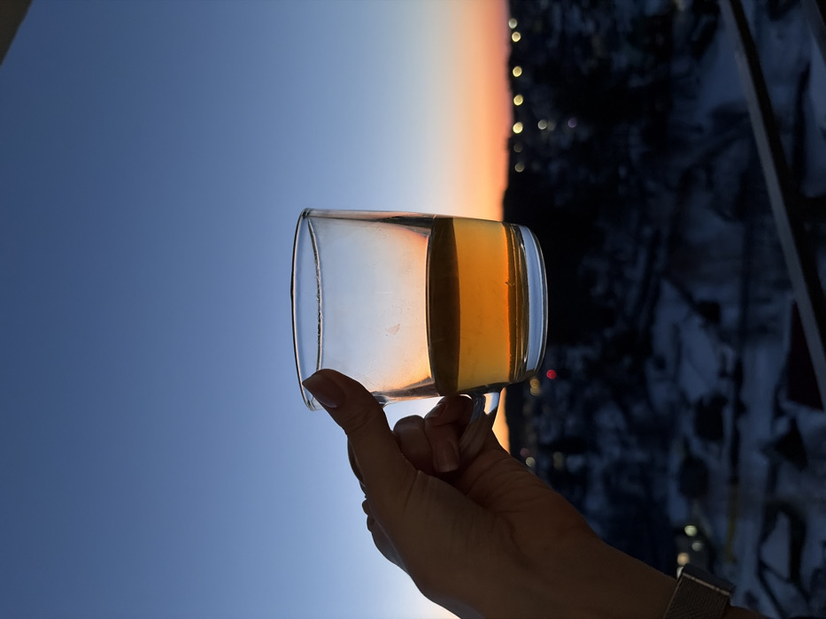 A right hand, displaying manicured nails and a silver mesh watch band on the wrist, holds a clear glass mug by its handle. The mug contains a golden-amber liquid, approximately half its capacity. The hand and mug are positioned against a twilight sky over Chaikovsky, Russia, which exhibits a gradient from deep blue overhead to an orange-pink hue at the horizon. Below the horizon, a blurred urban landscape is visible, characterized by dark structures, visible patches of snow, and numerous out-of-focus, bright lights. A dark, horizontal railing is partially visible at the bottom right of the frame, indicating an elevated vantage point.