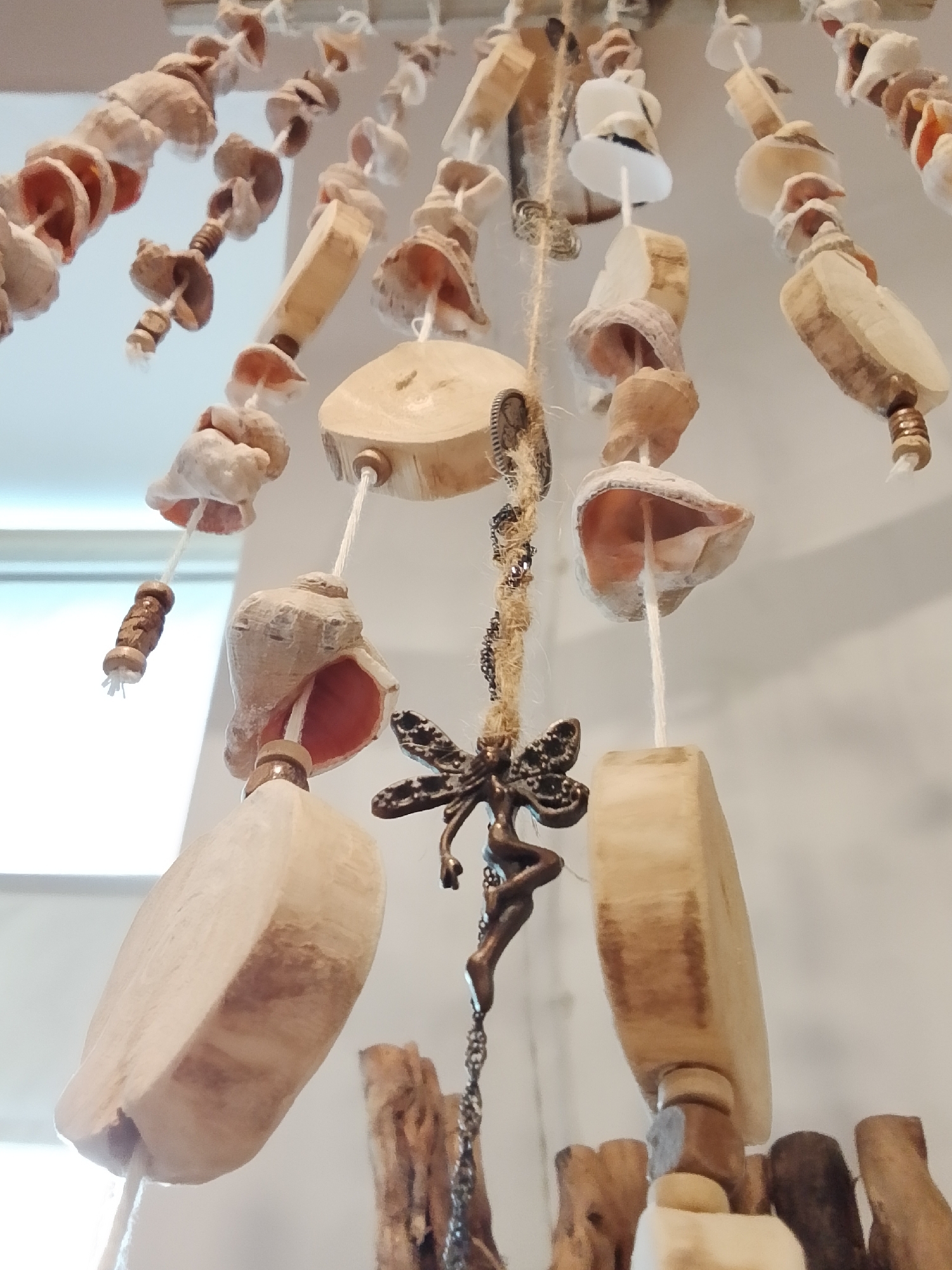 A low-angle, close-up shot focuses on a hanging wind chime. The wind chime consists of various materials including seashells, smooth wooden shapes, and a decorative fairy figurine suspended from chains. The hanging elements are strung from above, and the background includes parts of a white wall.
