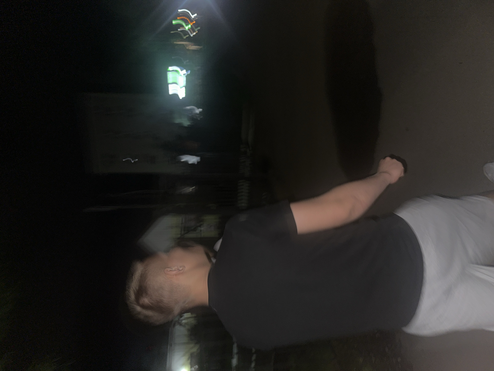 The image depicts a person in a dark, outdoor setting at night. The person is wearing a black t-shirt and light-colored pants. Their head is turned, and their hair is short and styled. One arm is raised, with the hand appearing clenched. The background is mostly dark, with some blurred lights visible in the upper left portion of the image, suggesting artificial illumination from buildings or vehicles. The overall impression is of a candid, possibly motion-blurred photograph taken in an urban environment at night. There is no visible text in the image. The location context provided is Birobidzhan, Russia.