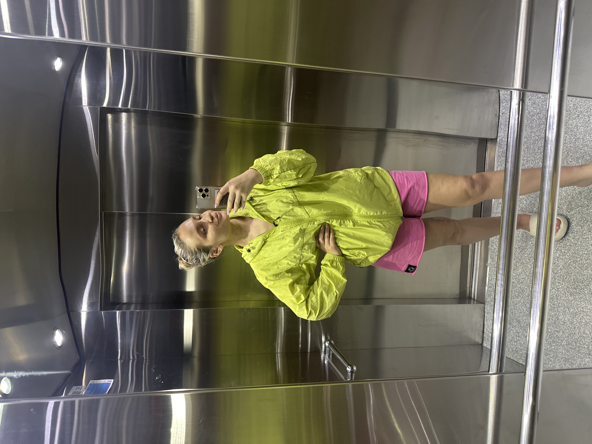 A woman is taking a selfie in what appears to be an elevator. The elevator has stainless steel walls and ceiling. She is wearing a light green jacket and pink shorts. Her hair is blond and pulled back. The elevator has a stainless steel handrail. The floor is speckled grey. There is no indication of the elevator's location in Fethiye, Turkey.