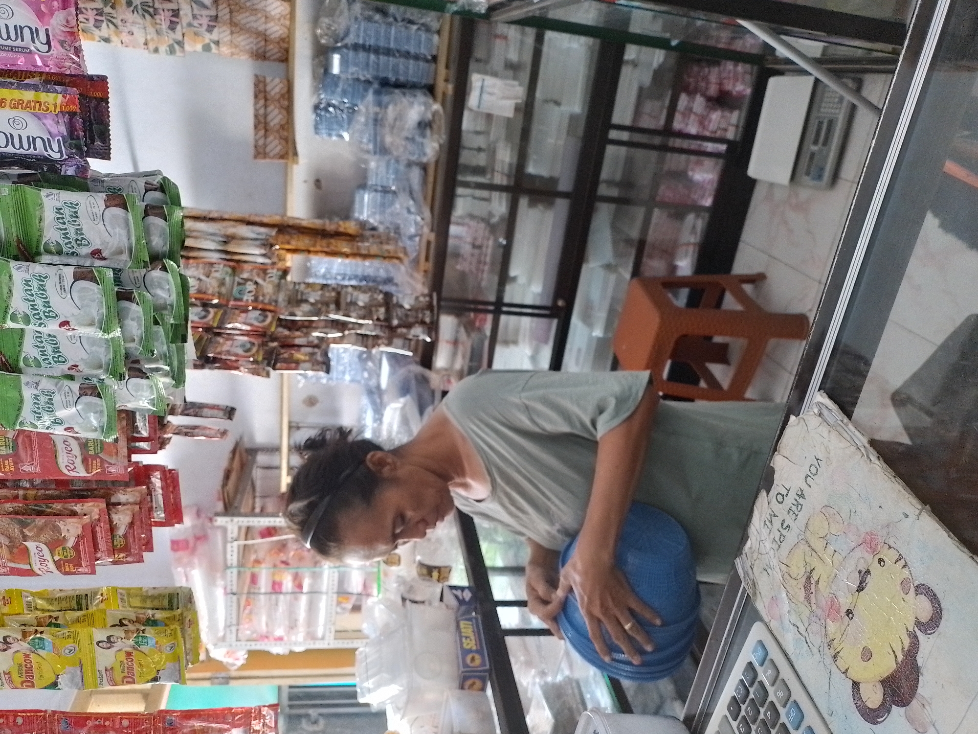An Indonesian woman is standing in what appears to be a small convenience store or warung in the city of Ngancar, Indonesia. She is wearing a green t-shirt and a black headband. She is holding a blue plastic bowl. In front of her is a calculator and a notebook with a cartoon drawing of a tiger and the text "YOU ARE SPECIAL TO ME". The store shelves are stocked with various packaged goods, including food items and plastic containers. A small orange stool is visible to her right. A digital scale is on the floor in the background.
