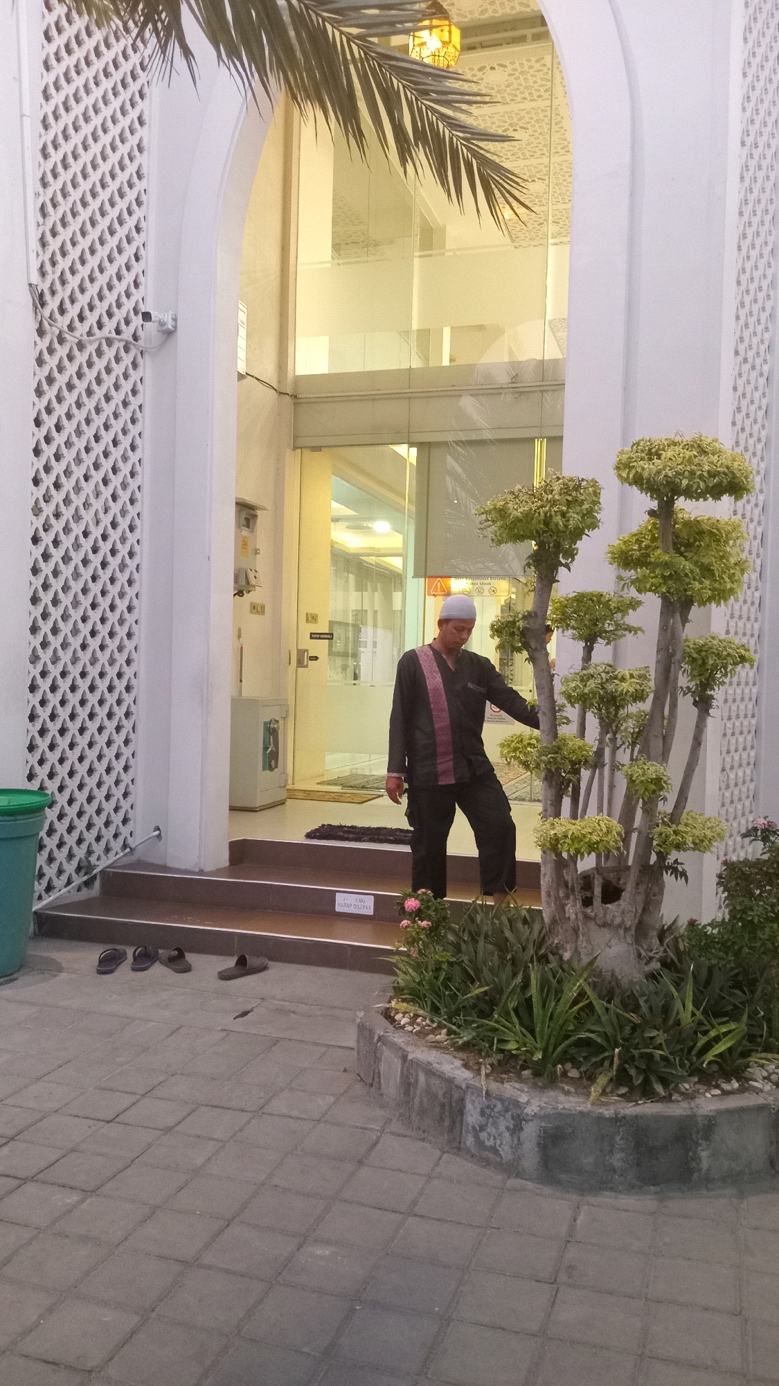 A man wearing a white kufi and a dark outfit with a pink stripe stands on a stepped entrance to a building. He is looking down and to his right. The building has white decorative panels on the left and right sides of the entrance. Inside the building, there is glass and lighting. To the left of the entrance, there is a green trash can. In front of the steps, there are several pairs of sandals. To the right of the entrance, there is a decorative tree with multiple branches and green foliage. The ground in front of the entrance is paved with square tiles. A sign with writing in Indonesian is visible on the steps.