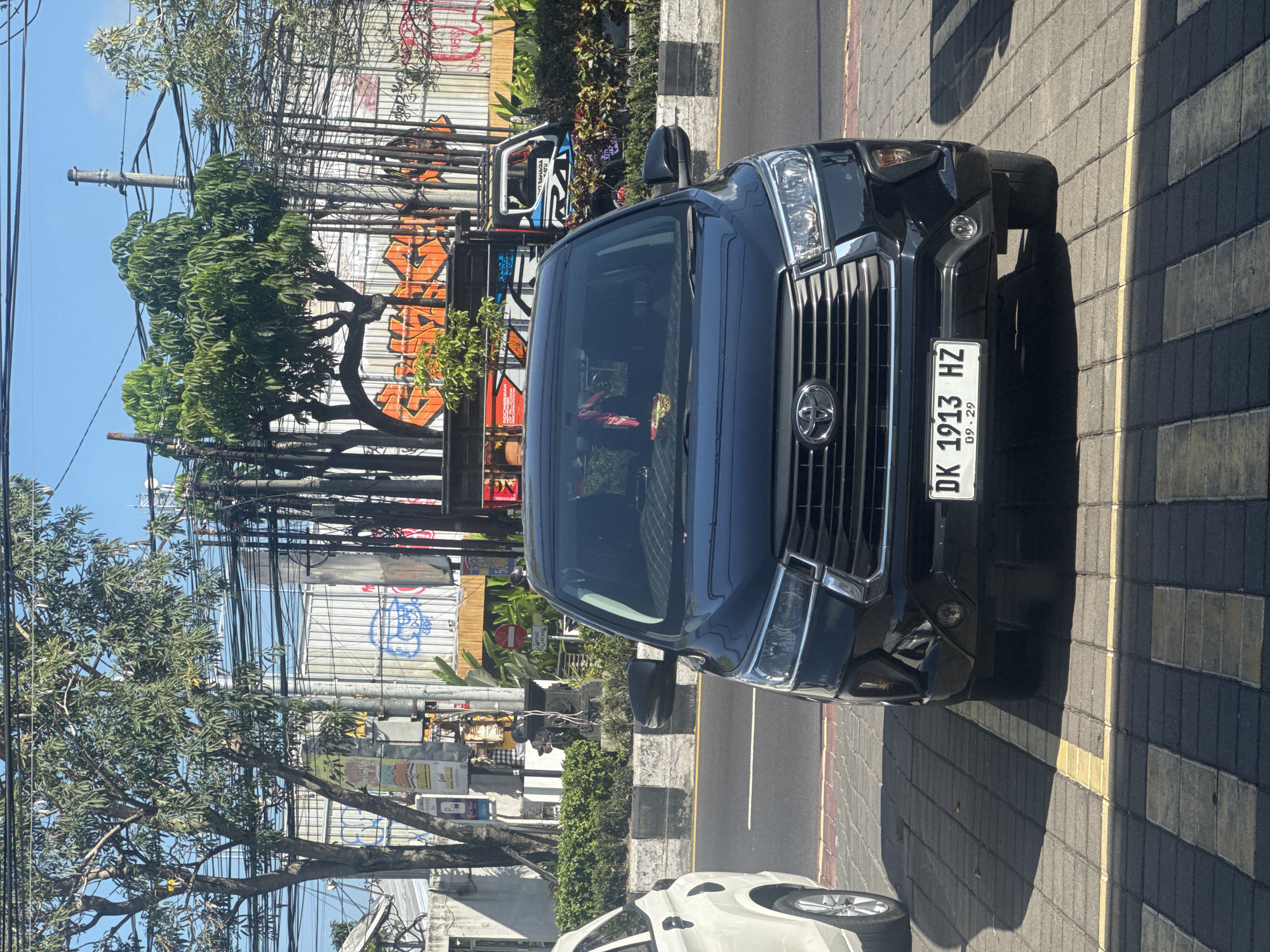 A black Toyota Innova is parked on a brick sidewalk in Kuta, Indonesia, with a white car partially visible to its left. The Innova has a license plate reading "DK 1913 HZ" and "09.29". Behind the cars, a street runs parallel to the sidewalk, separated by a curb. Further back, there is a wall covered in graffiti and various trees and utility poles with numerous wires. The sky is clear and blue.