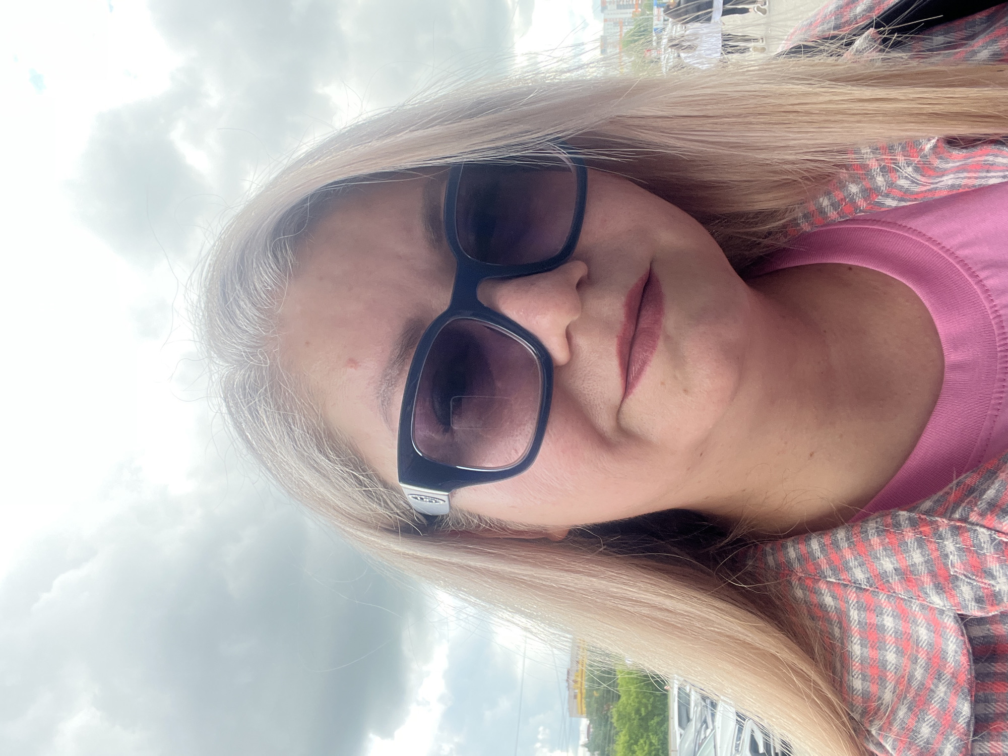 A medium, canted, selfie-style shot of a woman with long, straight, light blonde hair wearing dark-framed sunglasses, a pink t-shirt, and a red, gray, and white plaid flannel shirt. She is outside, with a cloudy sky visible above and in the background. In the distance, there are trees and buildings. Based on visual cues (likely the style of buildings and cars), the location may be Izhevsk, Russia.