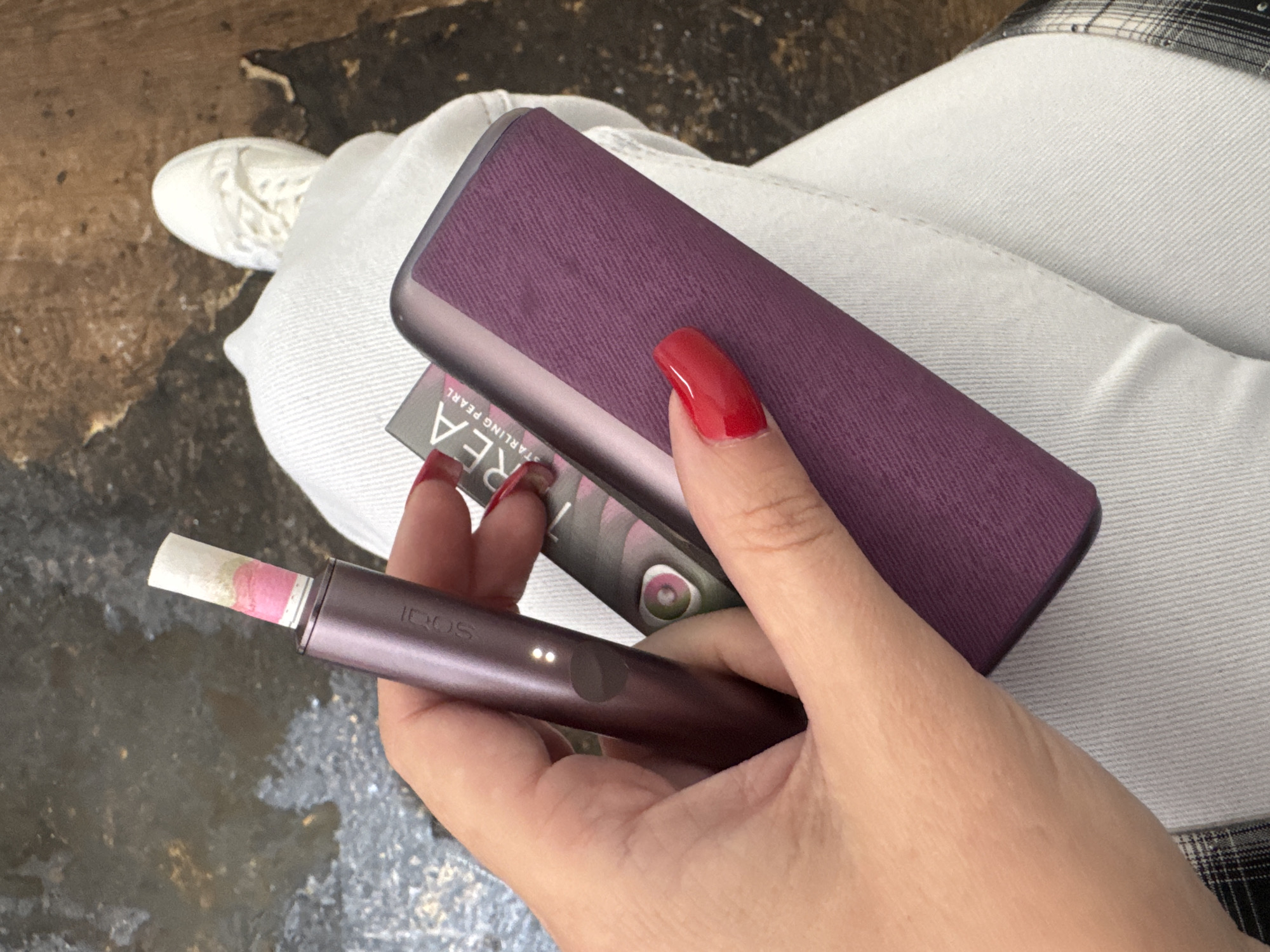 A woman's hand with bright red fingernails holds a purple IQOS electronic cigarette device, a pack of Terea cigarettes, and the charging case. The IQOS device has a light pink and white colored tobacco stick inserted. The charging case is also purple and has a textured surface. The woman is wearing white jeans and white sneakers. The background appears to be a floor with some textured and wet areas, possibly outdoors or in an industrial setting.