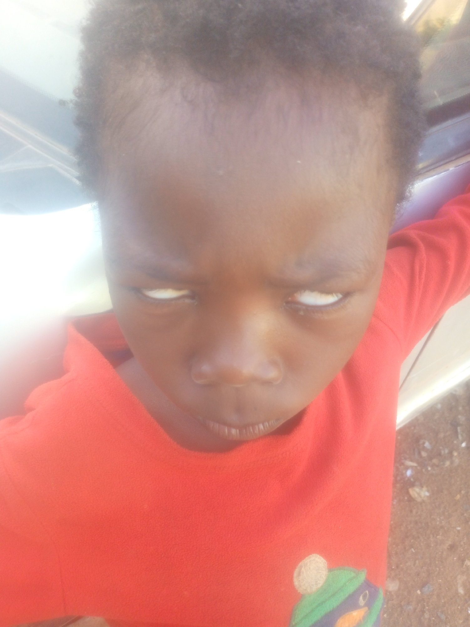 A close-up, high-angle portrait shows a young child with dark skin and short, dark curly hair. The child is wearing a bright red long-sleeved shirt, which features a partial graphic on the lower right corner depicting a cartoon-like character or animal wearing a green hat with blue and purple stripes, and a light-colored, round element above it. The child looks directly towards the camera, with both eyes rolled back to reveal only the whites, creating a distinctive and intense gaze. Their mouth is slightly parted. The scene is brightly lit, likely outdoors during the daytime, as indicated by the overexposed and washed-out areas in the background, particularly on the left side. Hints of a light-colored structure, possibly part of a vehicle, and blurry ground are visible in the background on the right. No discernible activity is taking place other than the child posing or reacting. There is no visible text.