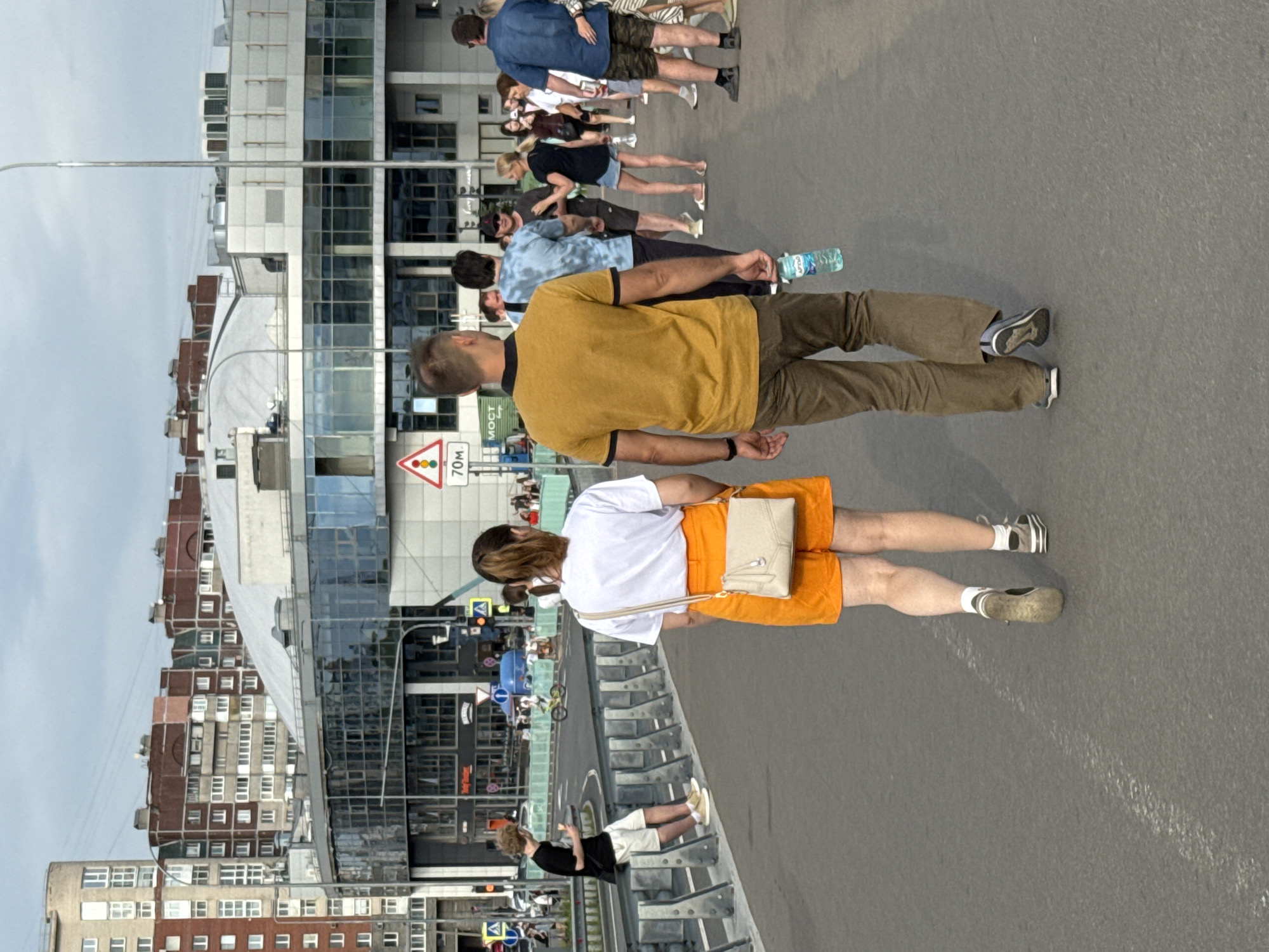 The image captures a street scene in Saint Petersburg, Russia, with several people walking along a paved road. The orientation of the image is sideways. In the foreground, a person wearing a yellow t-shirt and brown pants walks away from the camera. Behind them, a person in a white t-shirt and orange shorts is also walking away. Further back, a group of people are walking in the same direction, with some holding drinks.
The background features modern buildings with glass facades, characteristic of urban architecture. A traffic sign with "70M" is visible, indicating a distance marker. The sky is overcast, suggesting a cloudy day. The overall atmosphere appears to be casual, with people engaged in everyday activities like walking. The time of day is difficult to ascertain precisely due to the lighting conditions, but it seems to be daytime. No strong emotions are evident from the individuals captured in the frame.