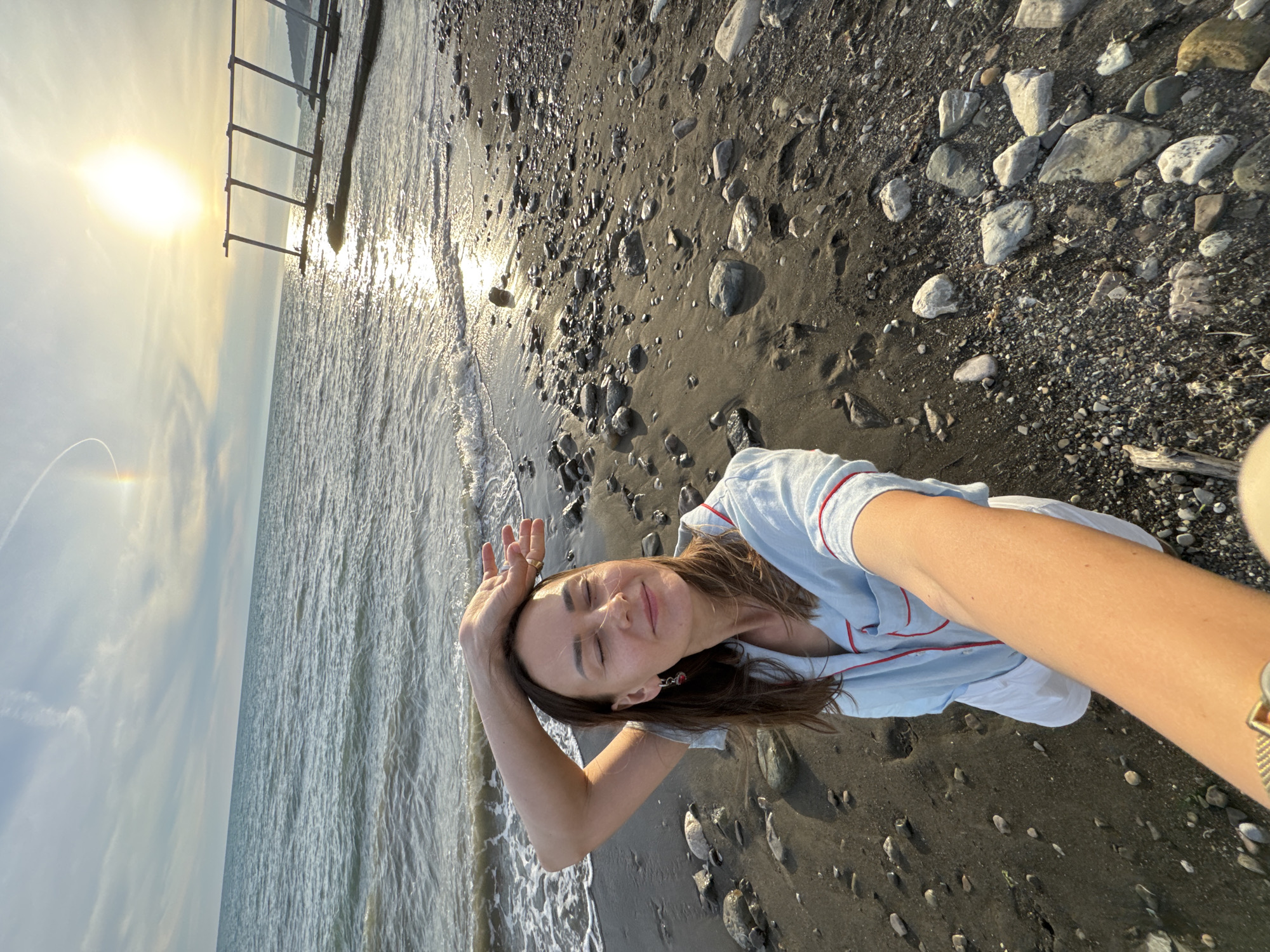 A woman is taking a selfie on a pebble beach, with the Black Sea behind her. She is wearing a light blue pajama set with red trim and white shorts. Her eyes are closed and her head is tilted back, with her right arm raised and bent behind her head. The sun is low in the sky, casting a warm glow and reflecting on the water. In the distance, there is a wooden pier structure extending into the sea. The beach is covered in dark sand and smooth, grey pebbles. The scene appears to be in Sochi, Russia, judging by the context provided.
