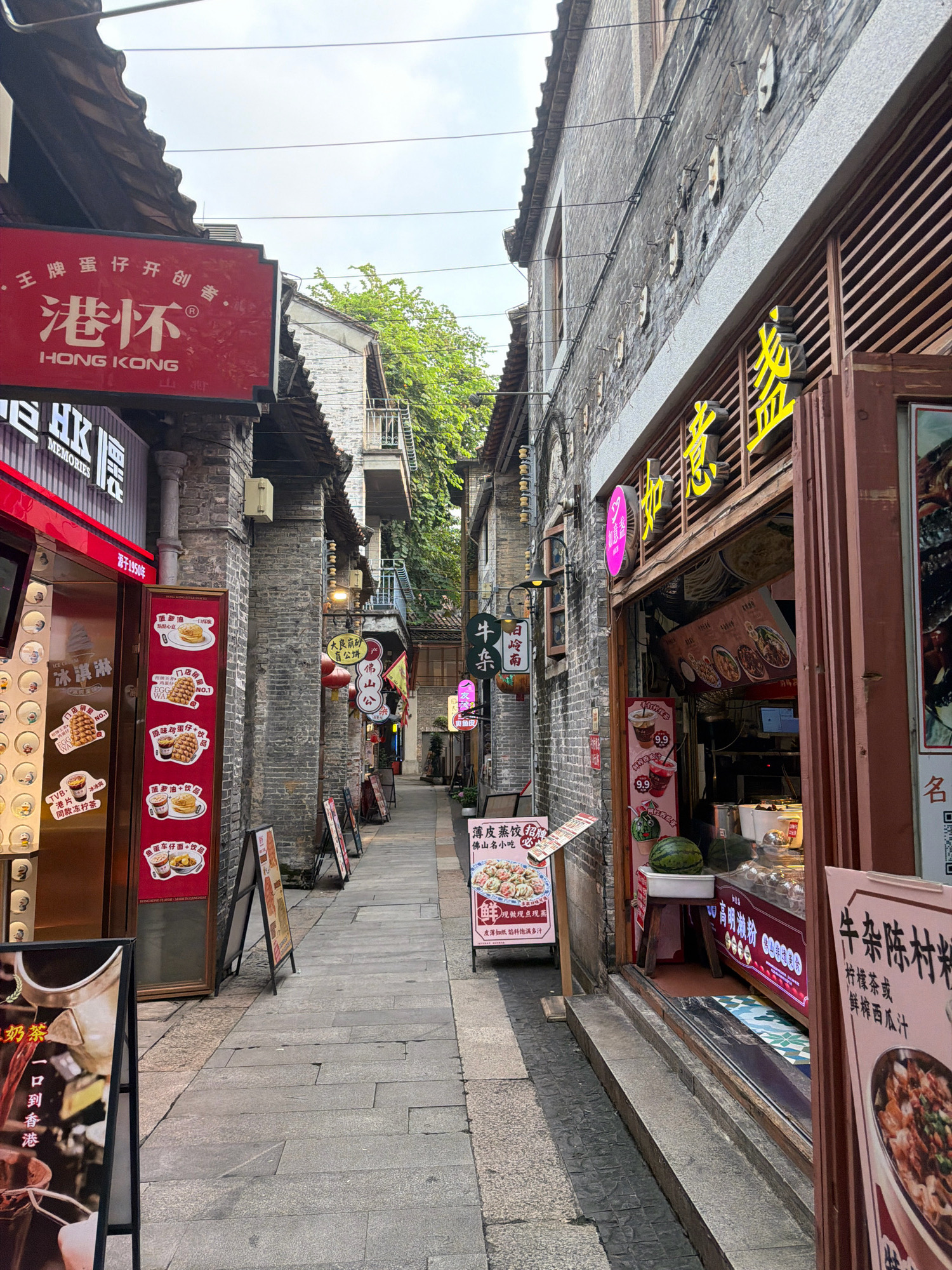 The image depicts a narrow alleyway in a bustling commercial district, likely in Chancheng District, China. The architecture is traditional, with grey brick buildings and dark tiled roofs. Shops line both sides of the alley, their storefronts adorned with vibrant signs in Chinese and some English.
On the left, a red sign with gold lettering announces "HONG KONG" and below it "MEMORIES," suggesting a Hong Kong-themed establishment. Another shop features a display of what appear to be food items and drinks, with images of waffles, pancakes, and beverages. Several A-frame signs stand on the sidewalk, advertising various food specials.
The right side of the alley also showcases shops with prominent signage. One shop has a large yellow sign with Chinese characters, and its entrance reveals a display of food items, including what looks like a watermelon. Another sign advertises "Beef Offal Noodles," and further down, a QR code is visible, hinting at modern payment methods.
The alley floor is paved with grey flagstones. The lighting suggests it is daytime, possibly late afternoon, with diffused sunlight filtering through the cloudy sky and the dense foliage of trees visible above the buildings. There are no people clearly visible in the scene, but the presence of shops and signage indicates a busy commercial area. The overall impression is one of a lively street filled with food vendors and shops, characteristic of a popular urban marketplace.