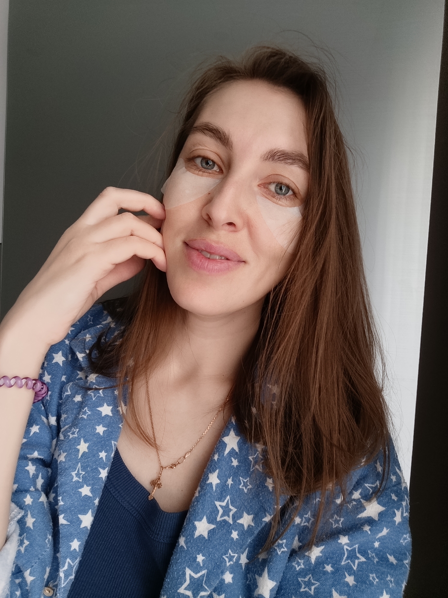 A woman is posing for a selfie indoors. She has fair skin, blue eyes, and long brown hair. She is wearing eye patches under her eyes. She's also wearing a blue shirt with white stars and a gold necklace with a cross. She has a purple bracelet on her left wrist and her left hand is touching her face. The background appears to be a simple wall, partially gray and partially white. The location is Chaikovsky, Russia.