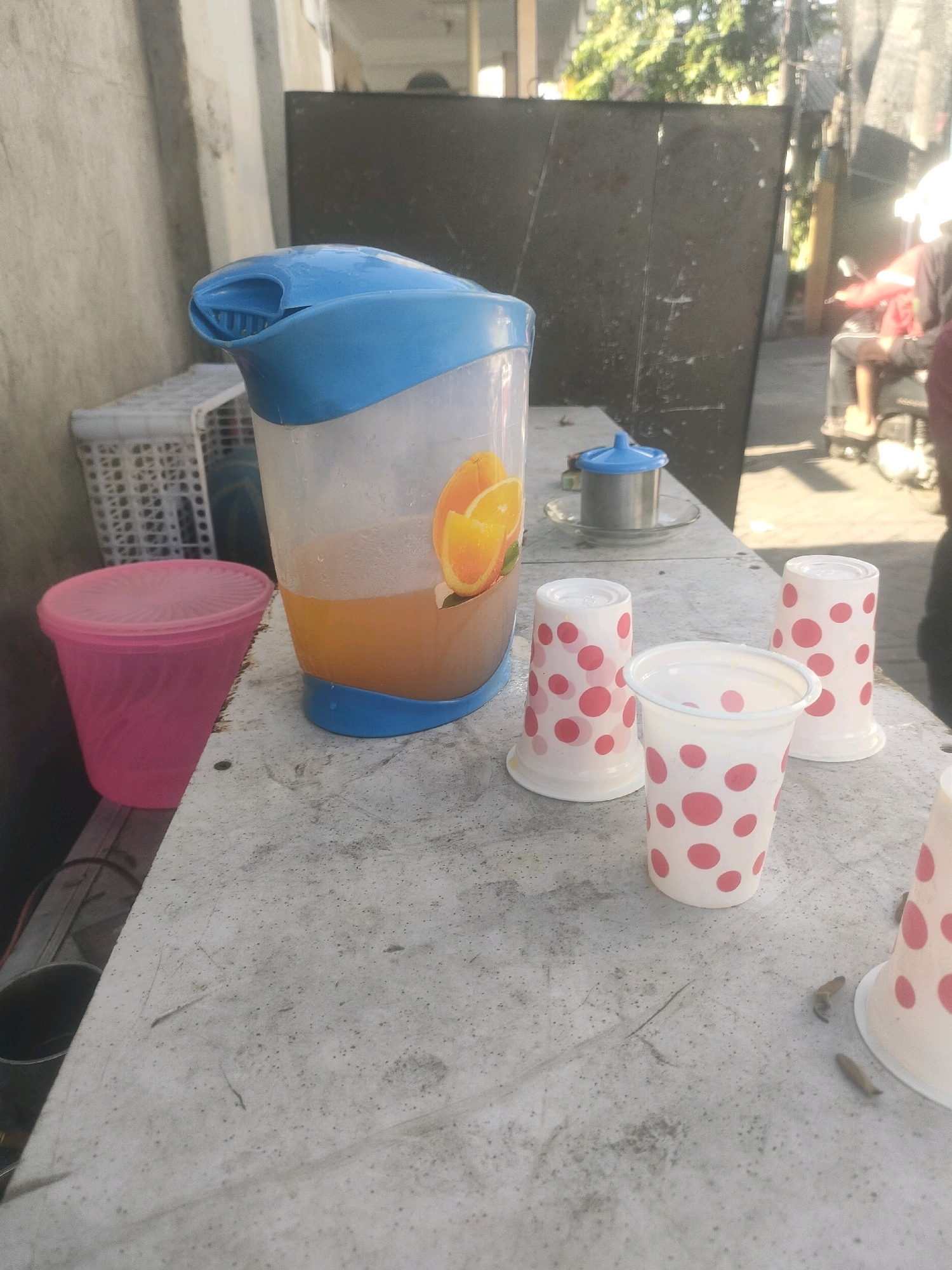 A close-up shot captures a pitcher of orange juice on a weathered, grey concrete table, likely in an outdoor setting in Surabaya, Indonesia. The pitcher, predominantly white with a blue lid and base, displays an orange slice graphic on its side. Partially visible next to it is a pink plastic container with a lid. Three paper cups adorned with pink polka dots are arranged on the table, one of which is upright and empty, while the other two are upside down. A small metal container with a blue lid rests behind the pitcher. In the background, a dark metal partition and blurred figures of people on motorcycles suggest a busy urban environment. Sunlight streams across the scene, casting faint shadows.