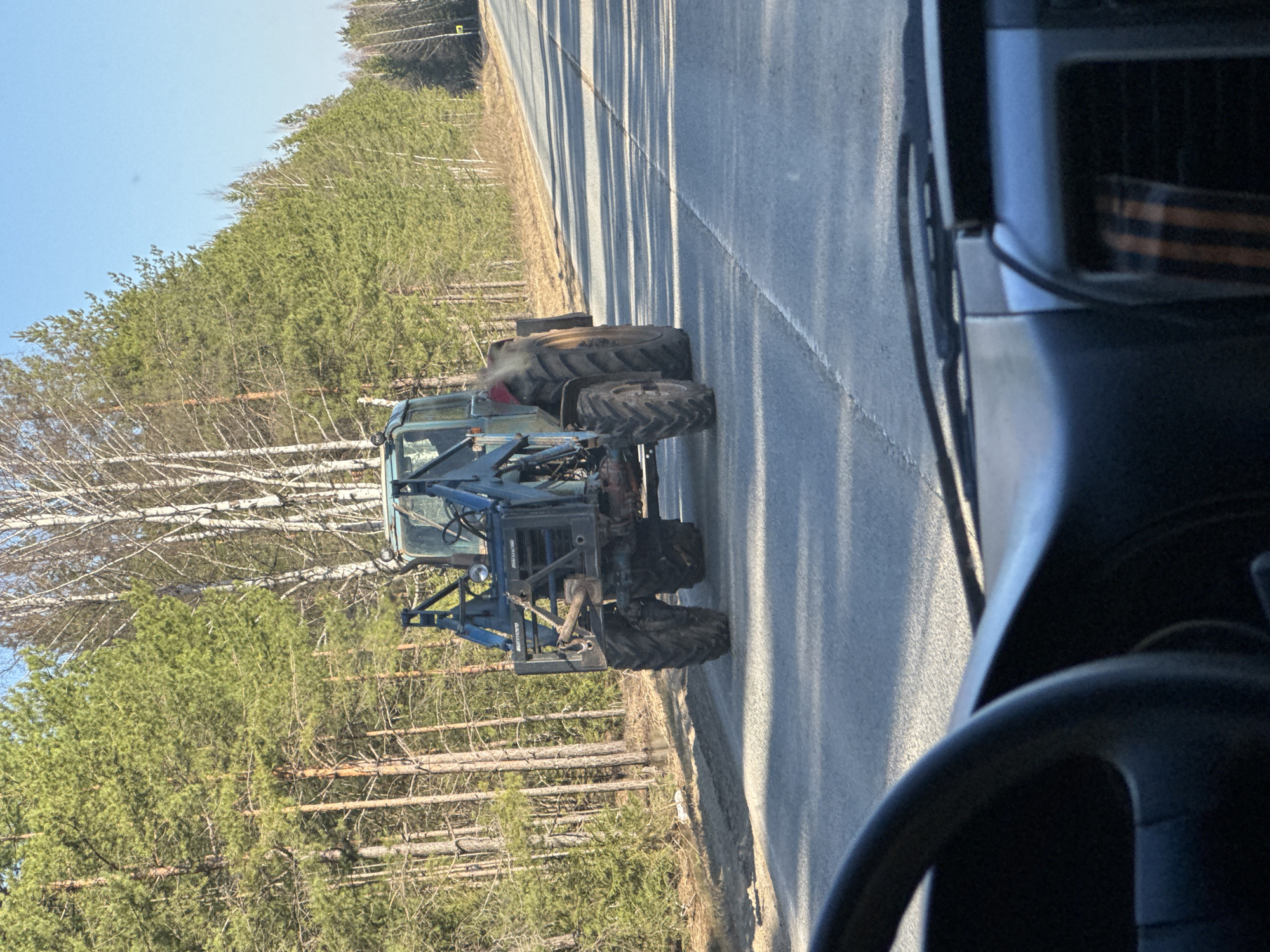 The image captures a rural outdoor scene, likely along a road in a forested area, with the location context provided as Krasnoyar, Russia.
The primary subject is a heavy-duty, blue or dark green forestry machine, resembling a forwarder or skidder, positioned on the shoulder of a paved road. It features large, deeply treaded tires indicative of off-road capability and rugged construction suitable for logging operations. The vehicle has a prominent hydraulic arm structure at its rear, although no logs are currently being carried. Visible text on the back of the machine appears to be "BROUTTE" and possibly "1010" or "1010T," suggesting it might be a Bruunett forestry vehicle.
The road is asphalt, with long, distinct shadows cast across it, hinting at either morning or late afternoon. The sky is clear and blue, indicating bright, sunny weather. To the left of the road, a dense forest stretches into the background. This forest comprises both coniferous trees, primarily pines with green foliage, and deciduous trees, likely birches, with their characteristic white bark and bare branches. Many felled logs are visible among the trees, reinforcing the forestry context of the scene.
The photograph is taken from inside another vehicle, with a portion of the dashboard and a steering wheel visible in the lower right foreground. Notably, a St. George ribbon, a symbol of military valor and patriotism in Russia, is affixed to the dashboard of the viewer's vehicle, providing a cultural detail. The activity appears to be the viewer driving past or stopped near the forestry machine, which itself might be parked or moving slowly.