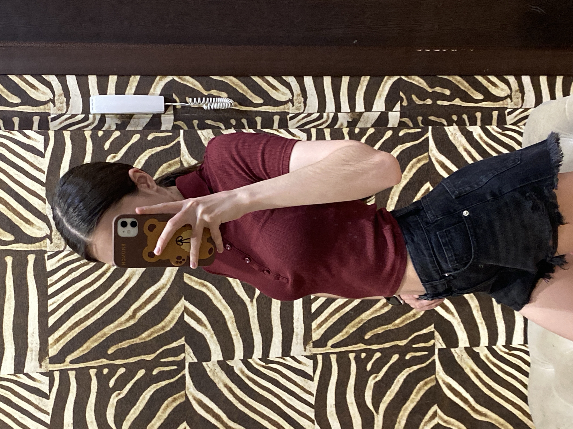 A person is lying on a bed covered with zebra-striped fabric, holding a phone with a brown bear case, and taking a selfie. The person is wearing a maroon collared shirt and black denim shorts. A white rectangular object with a coiled wire is on the bed near the head. The room has a dark brown headboard. There is no indication of the location being in Votkinsk, Russia.