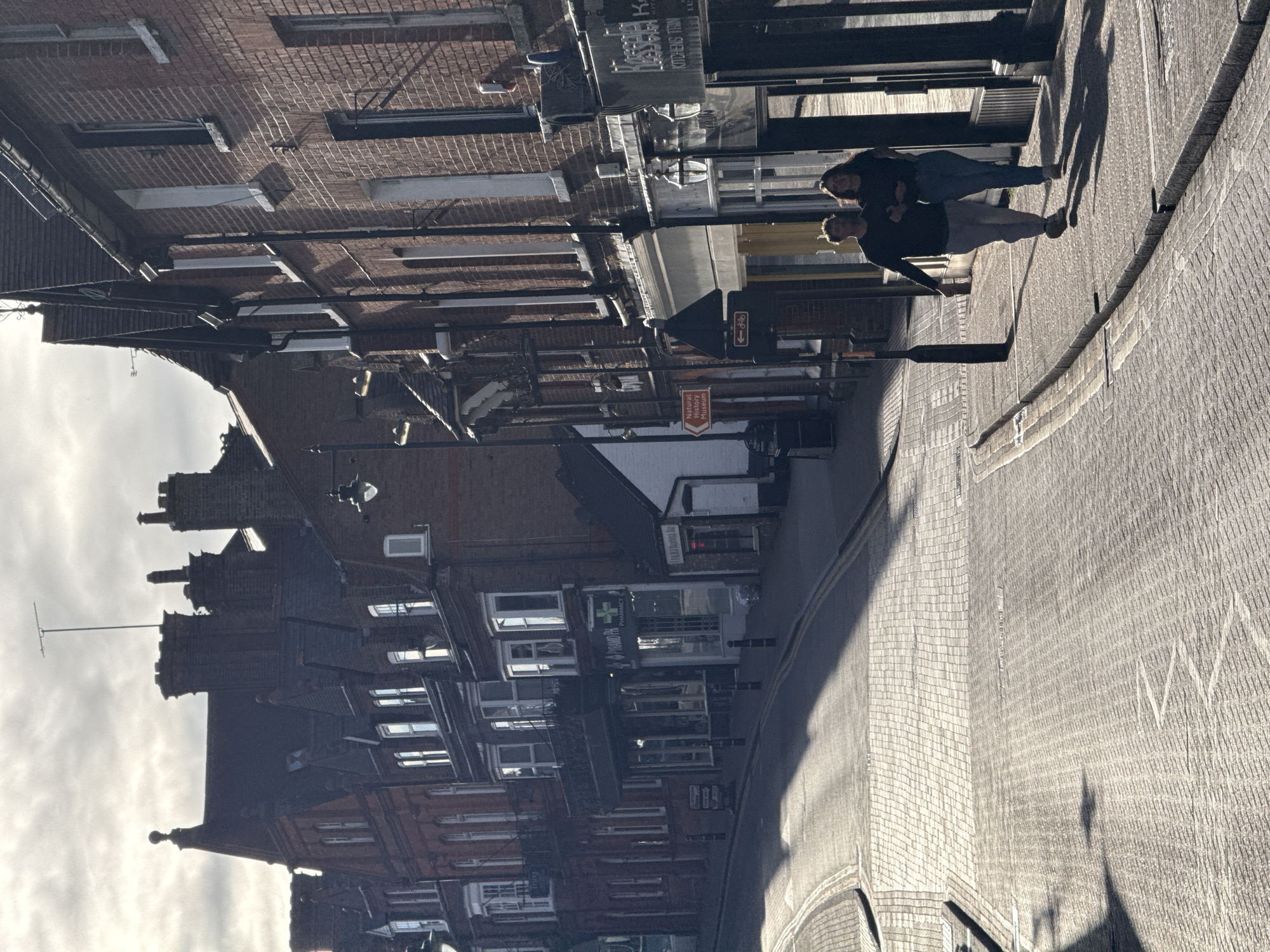 The image shows a street scene in Dacorum, United Kingdom. The street is lined with brick buildings of varying heights, featuring multiple windows and architectural details. On the right side of the frame, two individuals are walking along a paved pathway. Street signs and utility poles are visible along the buildings. The street surface appears to be paved with bricks. The sky is partially visible. A sign for the "Natural Museum" is visible on the building in the background. A pharmacy is on the building in the foreground.