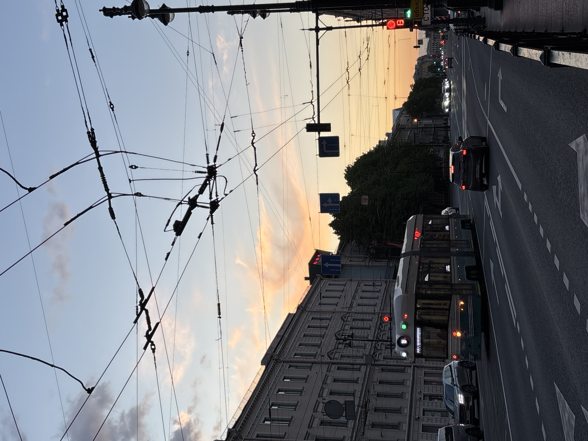 The image captures an urban street scene in Saint Petersburg, Russia, during either dawn or dusk. The sky presents a gradient of light blue, soft pink, and warm orange, indicative of the transitional lighting. A complex network of dark overhead electrical wires, typical for trolleybus infrastructure, dominates the upper part of the frame.
Below the wires, a multi-lane asphalt road stretches into the distance, marked with white dashed and solid lines, and white arrows indicating turns. Several vehicles are visible on the road, with their headlights and taillights illuminated. These include multiple cars and a large public transport vehicle, likely a trolleybus or bus, displaying red and white lights. A black SUV in the mid-ground clearly shows a Russian license plate "X070MC 178 RUS," confirming the Saint Petersburg region (178).
Multi-story buildings with numerous windows line the street, accompanied by some dark green tree foliage. Street furniture includes streetlights, traffic lights (some red, some green), and various signs. One traffic light structure displays green text, possibly "C TOP" or "СТОР," and another sign reads "ОБЪЕЗД" (Russian for detour). Two blue square traffic signs with white arrows indicate permissible movements: one shows a combined "straight ahead and right turn" and another a "left turn." A faint number "15" can also be seen on a pole near the sidewalk. The scene conveys a dynamic city environment at twilight.