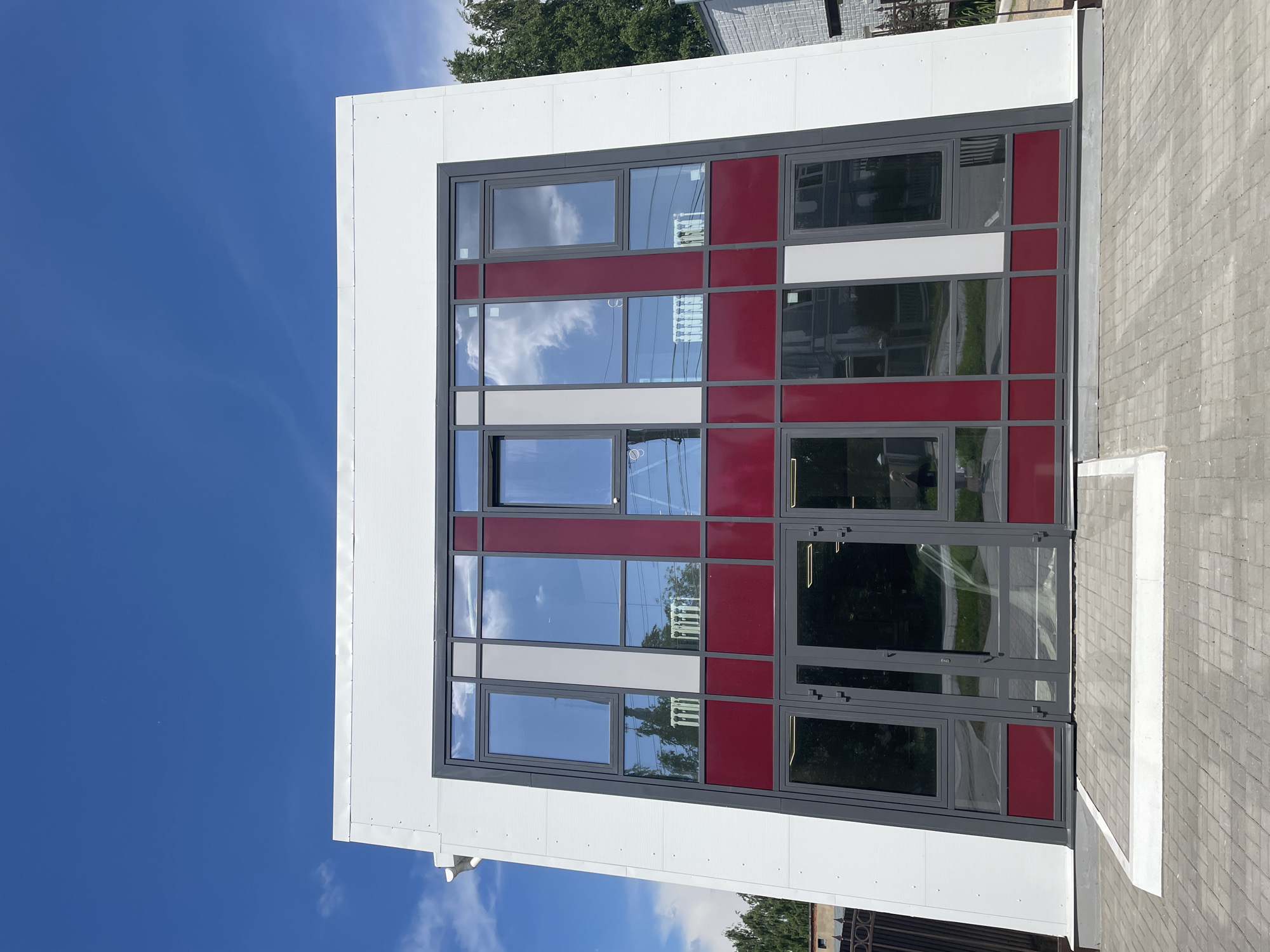 A modern, white, two-story building with a prominent glass facade dominates the image, set against a bright blue sky with scattered clouds. The facade features a grid of windows, some with a deep red accent, and others with a light grey or white panel. The windows reflect the sky and surrounding trees, as well as some interior elements that appear to be radiators. Below the main window area, there are double glass doors. The building is situated on a paved area, with a low, white concrete border in front. To the left, a dark metal fence is visible, and to the right, trees and parts of other buildings are seen. The scene appears to be a sunny day in Vologda, Russia.