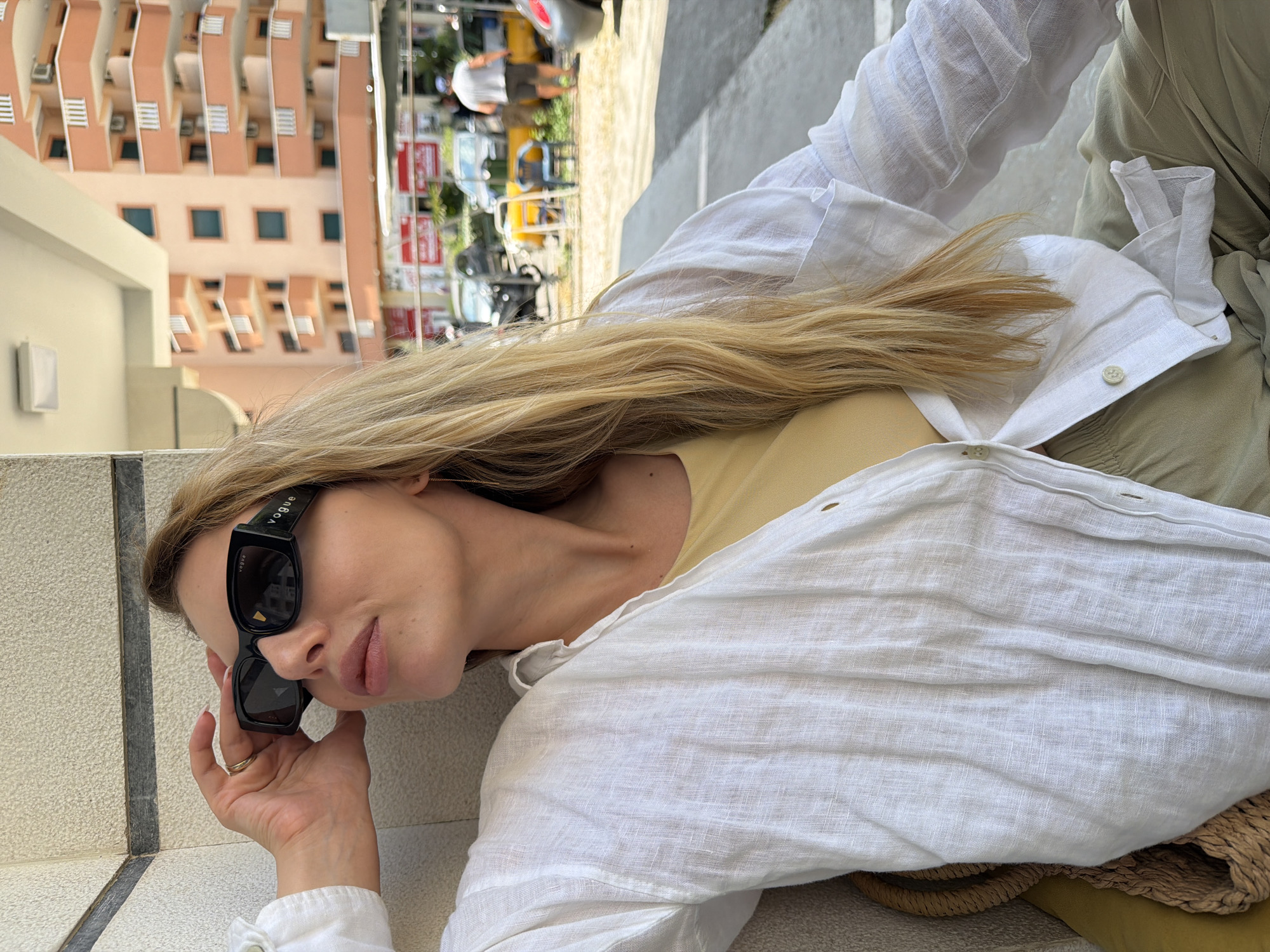 A woman with blonde hair, wearing large black Vogue sunglasses, a white linen shirt, and a light yellow tank top, is seated outdoors. She is looking up and to her left, with her right hand holding the side of her sunglasses. In the background, a peach-colored apartment building with white balconies is visible, suggesting an urban setting. Cars and people are also present in the distance. The scene appears to be in Budva, Montenegro, indicated by the architectural style and the overall atmosphere.