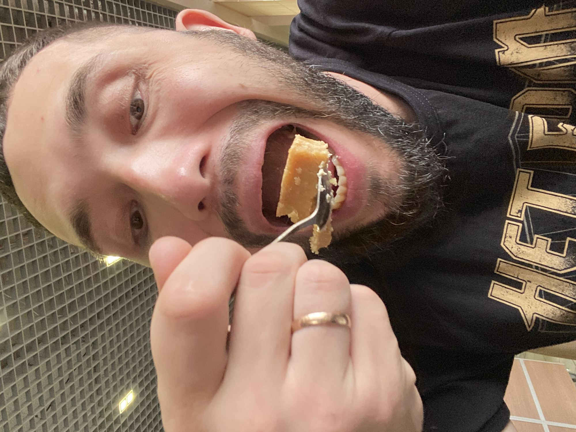 A close-up, tilted shot depicts a man with a light complexion, a dark beard, and brown eyes eating a piece of food with a fork. He is wearing a black t-shirt with a gold-colored design. A gold ring is visible on the ring finger of his hand holding the fork. The background appears to be a tiled wall. The lighting suggests an indoor environment. No evidence suggests the location is in Chaikovsky, Russia.