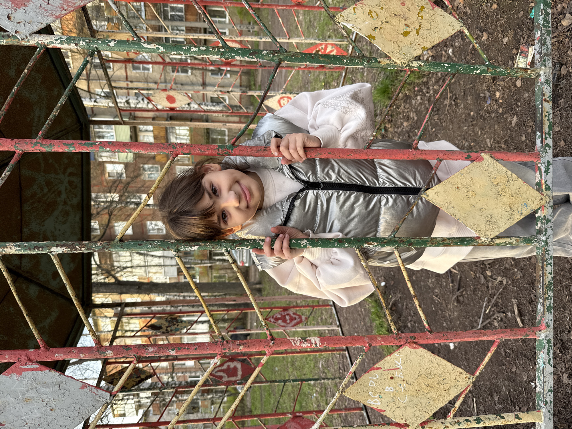 A young girl with brown hair and eyes, wearing a silver puffer jacket and white sweater, is holding onto an old, weathered metal fence. The fence has a diamond pattern and is painted with peeling layers of red, green, and yellow paint. Behind the fence, a multi-story apartment building is visible. The ground around the fence is covered with dirt and sparse patches of grass. The image appears to have been taken in Rusanovo, Russia.
