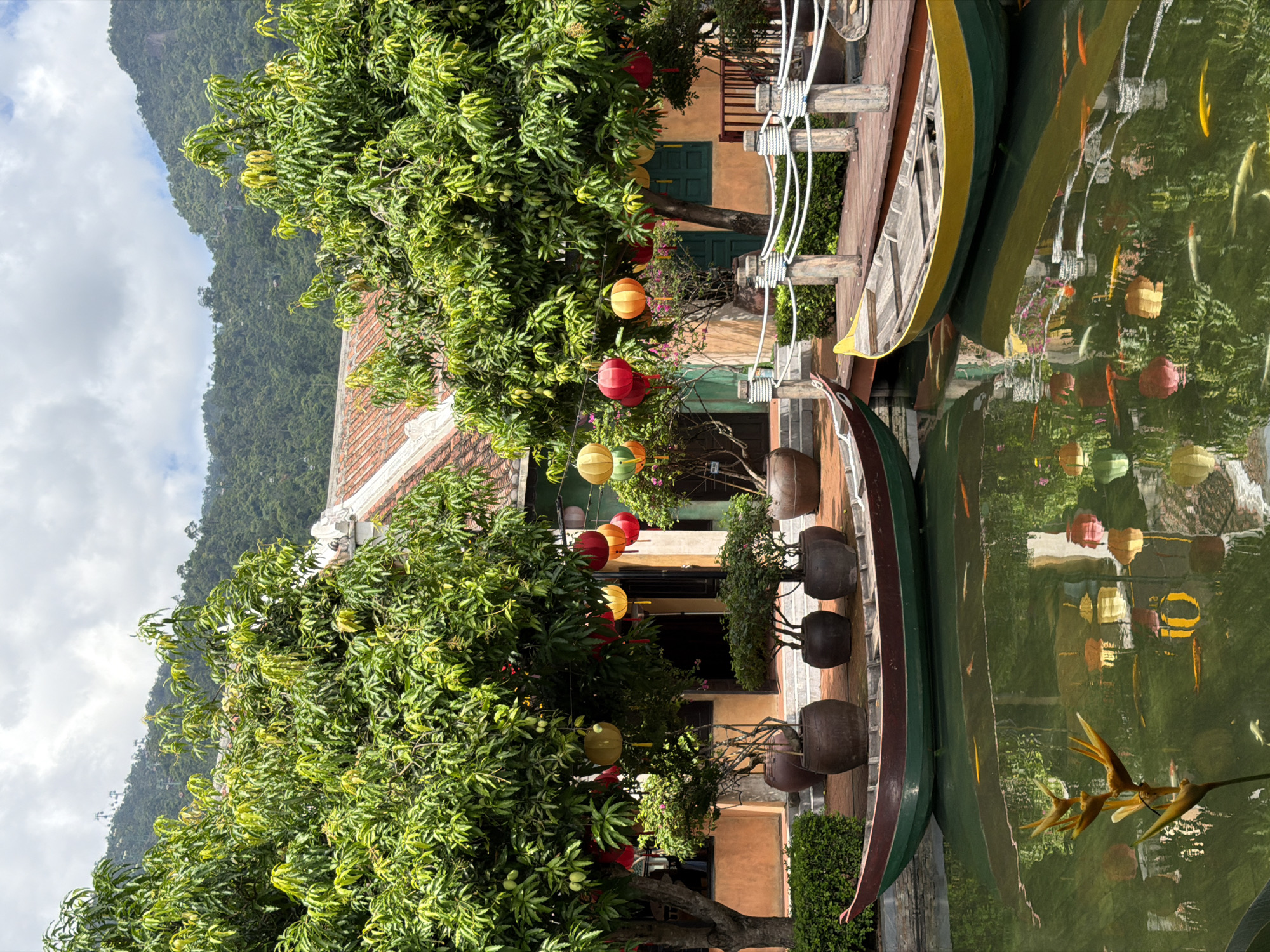 The image depicts a serene courtyard scene, likely part of a resort or cultural attraction in Da Nang, Vietnam, situated at the base of the Sơn Trà Peninsula. The focal point is a tranquil pond reflecting lush greenery, colorful lanterns, and the surrounding architecture. Two traditional wooden boats, one with a red and green hull and the other with a yellow and green hull, are moored at the edge of the pond. The pond is populated with numerous koi fish, their vibrant scales creating streaks of orange and gold in the water.
In the background, a traditional Vietnamese building with an orange facade and a terracotta tiled roof stands partially obscured by mature trees. These trees are adorned with various colorful lanterns, including red, yellow, and green, which hang from their branches. Potted plants and trees are strategically placed on a paved area bordering the pond, contributing to the overall aesthetic.
A wooden walkway, secured by sturdy posts and white ropes, runs along the edge of the pond, providing a pathway. Beyond the courtyard, the dense, verdant slopes of the Sơn Trà Peninsula rise, shrouded in a light haze and under a partially cloudy sky. Cable cars are visible ascending the mountainside, hinting at the presence of a cable car system, potentially leading to tourist attractions like the Golden Bridge or Ba Na Hills.
The scene is devoid of people, creating a sense of peaceful solitude. The natural elements of the trees, water, and mountains are complemented by the cultural elements of the traditional architecture and lanterns, suggesting a place designed for relaxation and cultural appreciation. The lighting indicates it is daytime, with diffused sunlight filtering through the clouds.