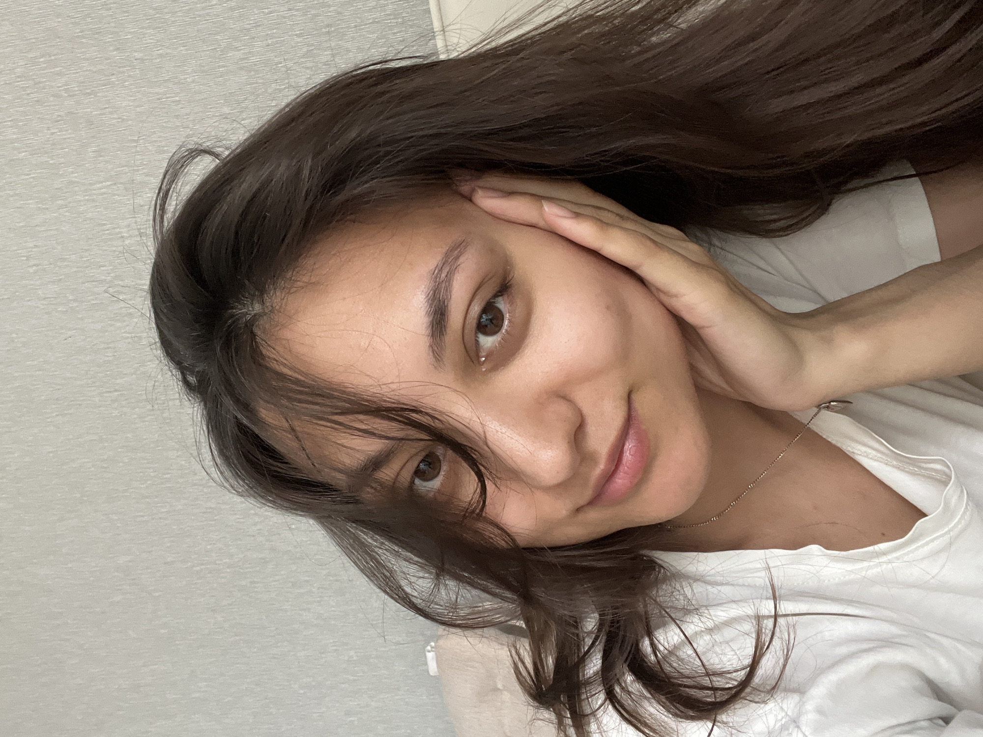 A close-up, selfie-style portrait shows a young woman with dark hair and light brown eyes. Her hair falls around her face, partially obscuring her left eye. She is wearing a white t-shirt and a delicate gold necklace. Her left hand is gently touching her cheek. The background appears to be a light grey or white wall. There is no information provided in the image about the location being Yekaterinburg, Russia. The description is limited to what is visually discernible.