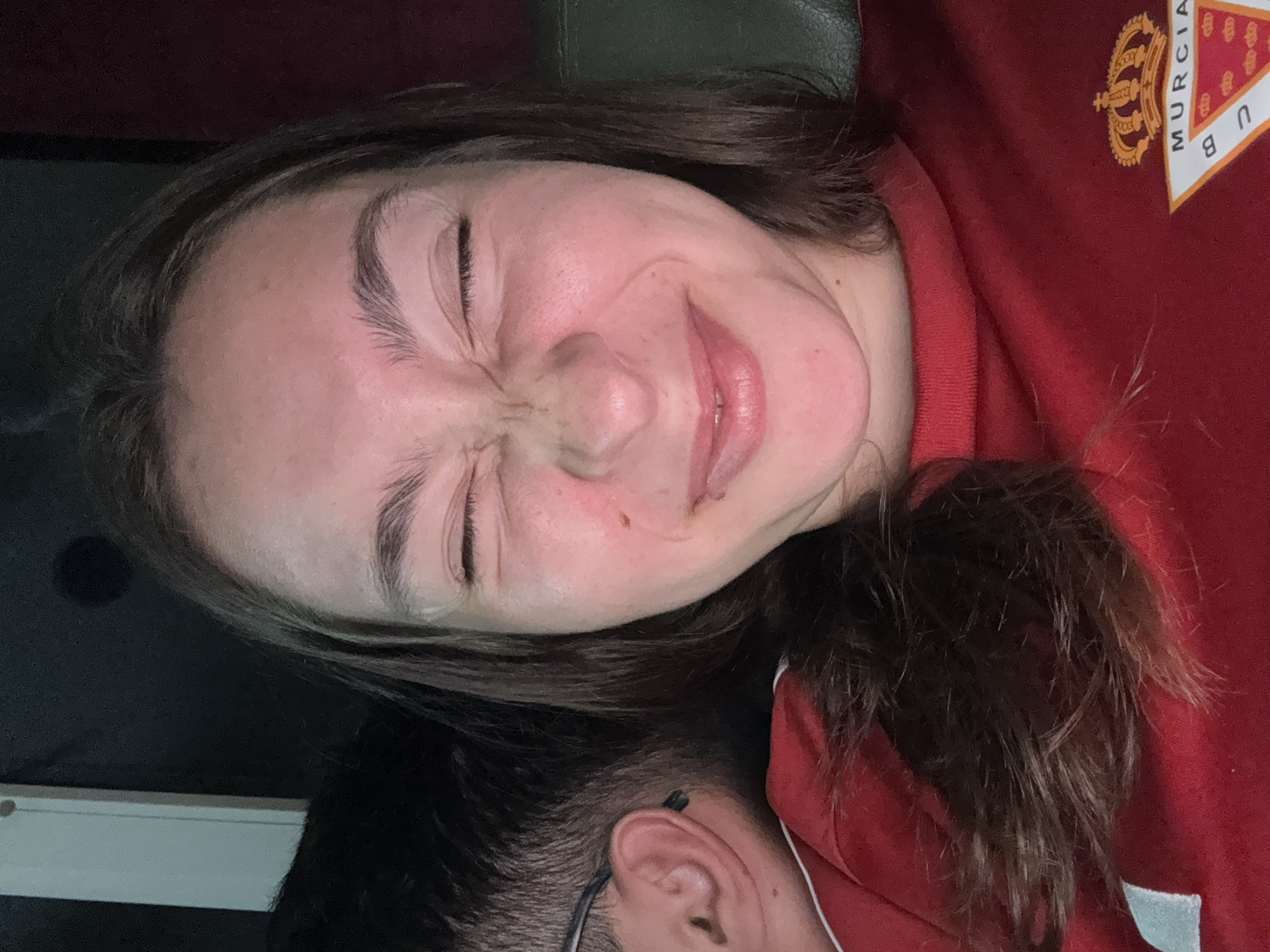 A close-up, head-and-shoulders, selfie-style photograph shows a young woman with brown hair and her eyes squeezed shut, making a playful grimace. Her face is illuminated, revealing a light complexion with some redness on her cheeks. She is wearing a red shirt with a crest visible on the lower right side of the frame. Partially visible behind her, to the left, is the side profile of a person wearing glasses and a black shirt. The background is dark, with a hint of what appears to be a light-colored vertical object on the far left. The lighting suggests an indoor setting at night, possibly in a private residence in Sofia, Bulgaria, given the absence of any identifiable landmarks or urban context.