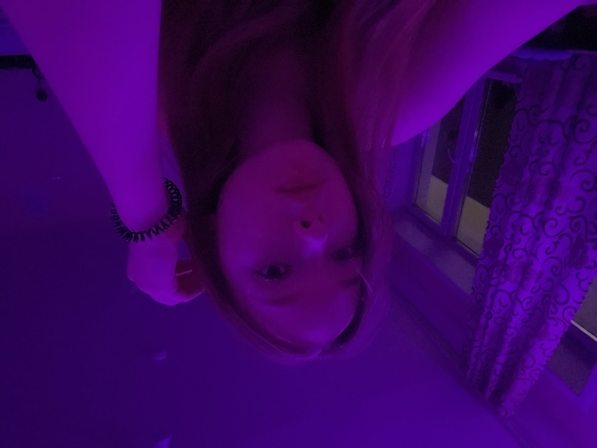A person is seen in an indoor setting illuminated by purple lighting. The location could potentially be in Chaikovsky, Russia, but there is no way to verify this information. The person is facing the camera at an angle, with their head tilted. The person has long hair. The arm is visible, with a bracelet or hair tie around the wrist. In the background, there is a window with curtains with a patterned design.