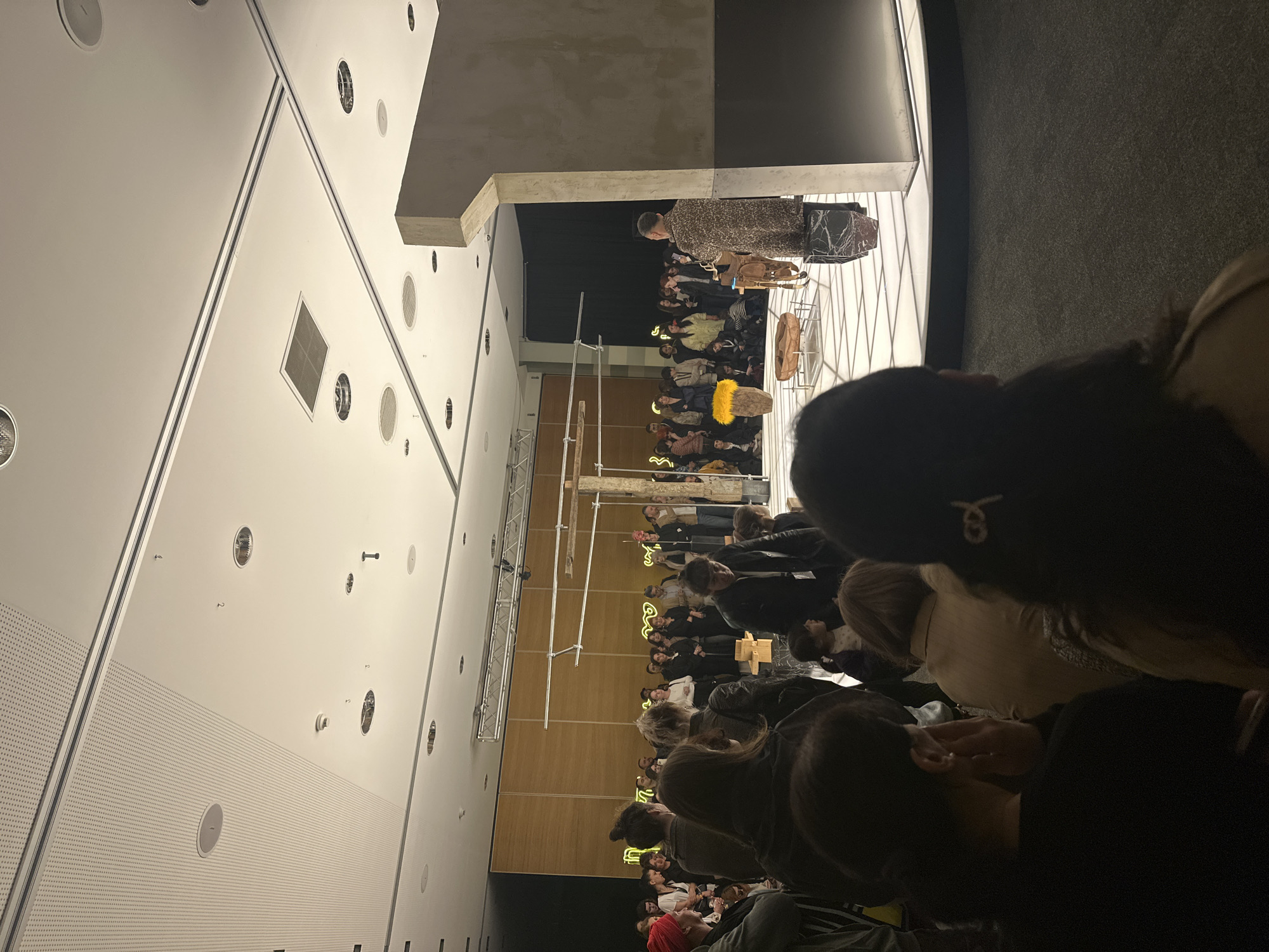 The image captures a fashion show or presentation in what appears to be an indoor venue in Tbilisi, Georgia. Attendees are seated in rows, observing models or performers on a platform or stage. The venue has a modern, minimalist aesthetic, with a high white ceiling, perforated panels, and numerous circular light fixtures. Some scaffolding is visible. Several people are in focus in the foreground, seated and observing the scene, while the stage is occupied by a person and several objects or displays. The overall lighting is dim.