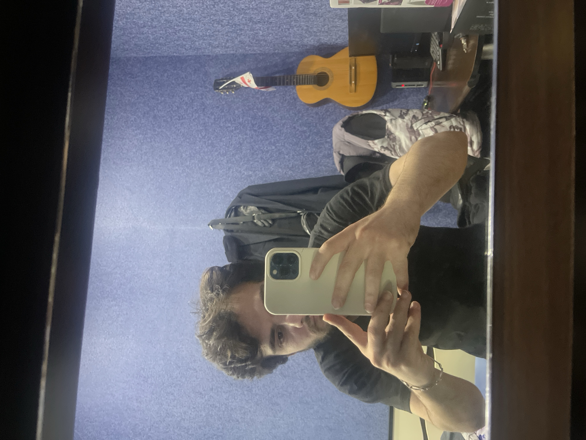 A young man with dark, curly hair and a beard is taking a mirror selfie in a room. He is wearing a black t-shirt and a silver bracelet on his left wrist. He is holding a light-colored smartphone in his hands, with which he is capturing the reflection.
The room has blue textured walls. To the right of the man, a light brown acoustic guitar is leaning against the wall, adorned with a small flag of Georgia. Behind the man, on a hook, is a dark jacket. Further to the right, a desk is visible with a computer monitor, keyboard, and other miscellaneous items. The mirror reflects the man and the objects in the room. The overall scene suggests a personal space, possibly a home or a studio, located in Tbilisi, Georgia, as indicated by the flag.