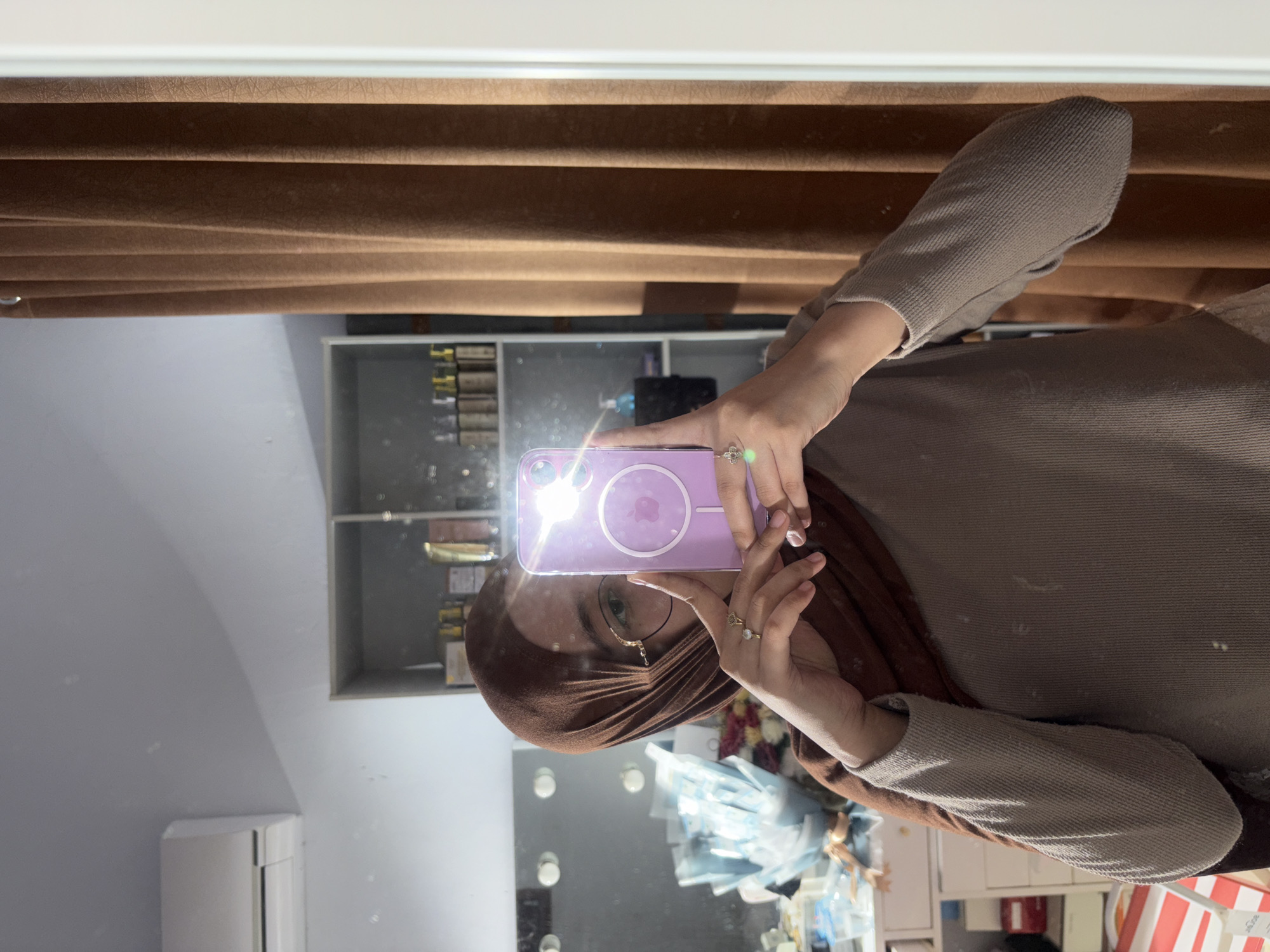 A person wearing a brown hijab, eyeglasses, and a gray top is taking a mirror selfie with a purple phone. The reflection in the mirror shows a room with a brown curtain, a shelf with various items, an air conditioner, a mirror with multiple light bulbs, and an assortment of objects. The location is presumed to be Gadingrejo, Indonesia.