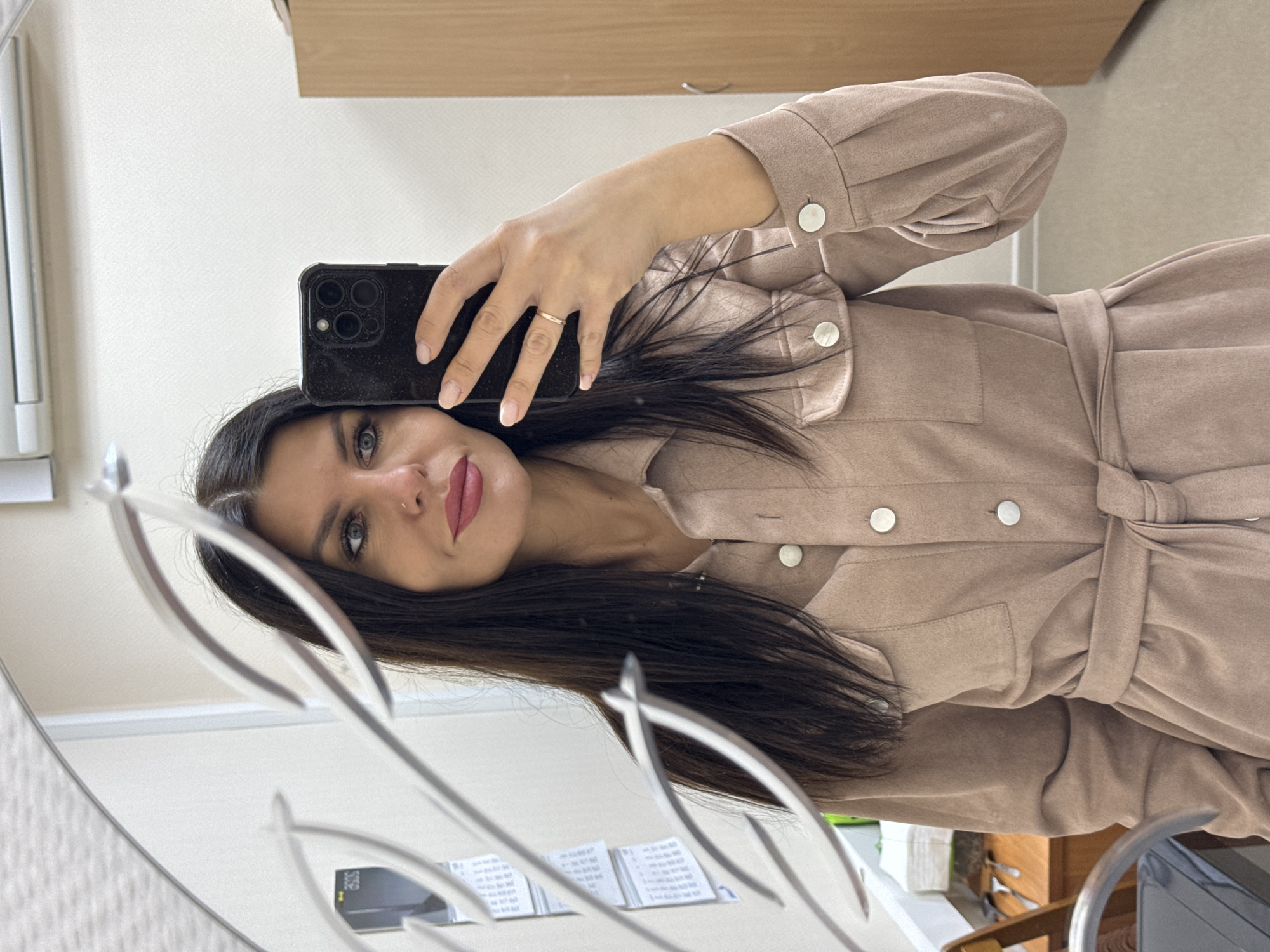 The image, rotated 90 degrees clockwise, depicts a mirror selfie of a woman with long, dark hair and blue-green eyes. She wears dark pink lipstick and has a small nose piercing. Her expression is a soft, neutral smile. She is dressed in a light beige or tan collared garment, which appears to be a dress or a shacket, featuring a matching tie-belt cinched at the waist and round, silver-colored buttons.
She holds a dark smartphone, identifiable by its triple camera lenses on the back, in her left hand to capture the picture. A simple gold-colored ring is visible on her left ring finger, and her nails are painted with a light pink or nude polish.
The mirror's reflective surface is prominent, with a decorative, abstract silver-colored frame or edge visible in the foreground. The background, seen as a reflection in the mirror, features a plain light-colored wall with a subtle textured pattern. A light brown wooden cabinet or shelf is reflected above her head. On a surface below her, several small white papers or cards with illegible text are visible, along with a faint "303S" on one reflected object. The indoor setting suggests an office, dressing room, or a well-lit room in a home. The time of day and weather are not discernible from the image.