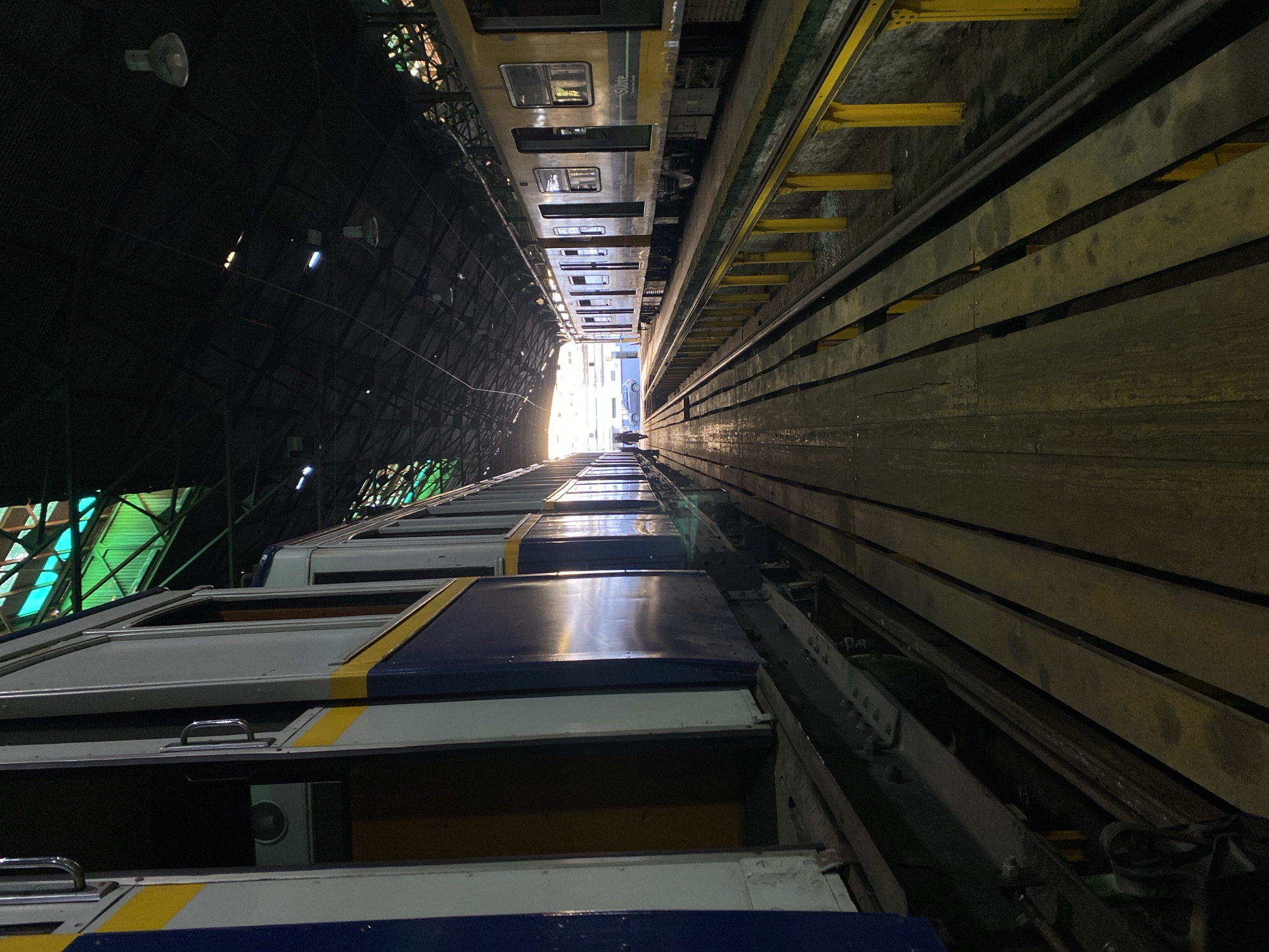 The image depicts the interior of a train depot or workshop, likely located in Buenos Aires, Argentina, as the subway car visible has a "Subte" (short for "Subterráneo," meaning subway in Spanish) logo, which is a term used to reference the subway system in that city.
Several train cars are lined up, with two lines of cars on either side of the image. The sides of the cars display a blue and white colour scheme. Along the right-hand side, a wooden walkway or platform runs parallel to the trains, supported by yellow steel or iron beams.
The ceiling is high and dark, with visible support structures and some lighting fixtures. Natural light streams in from the end of the depot, illuminating the far end of the train tracks.
There are no visible people in the image. The scene suggests a maintenance or storage facility for subway cars. The viewpoint is unusual, which adds to the feeling of it being an unusual angle, looking up or down an incredibly tall space, perhaps making the viewer feel disoriented.