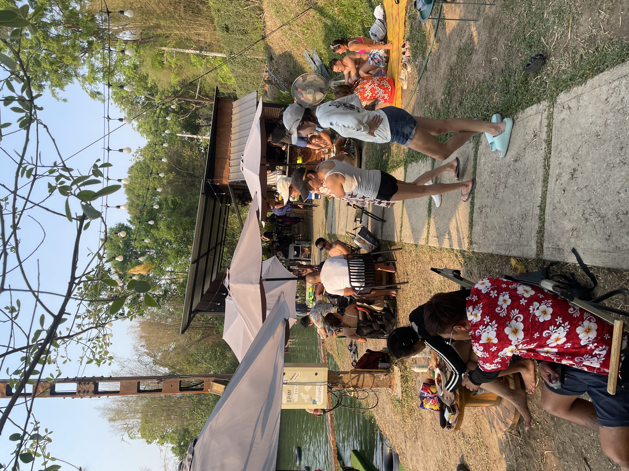 This image captures a lakeside scene in Tambon Nam Phrae, Thailand. Several people are engaged in different activities. Some individuals are seated under umbrellas, appearing to be dining or relaxing. Others are sunbathing on the grass, while a couple is walking away from the main group. An outdoor cafe or restaurant is present, with tables and chairs set up for patrons. A body of water is visible, with some form of electric equipment located near the shoreline. The environment includes lush greenery and trees, indicating a natural setting.
