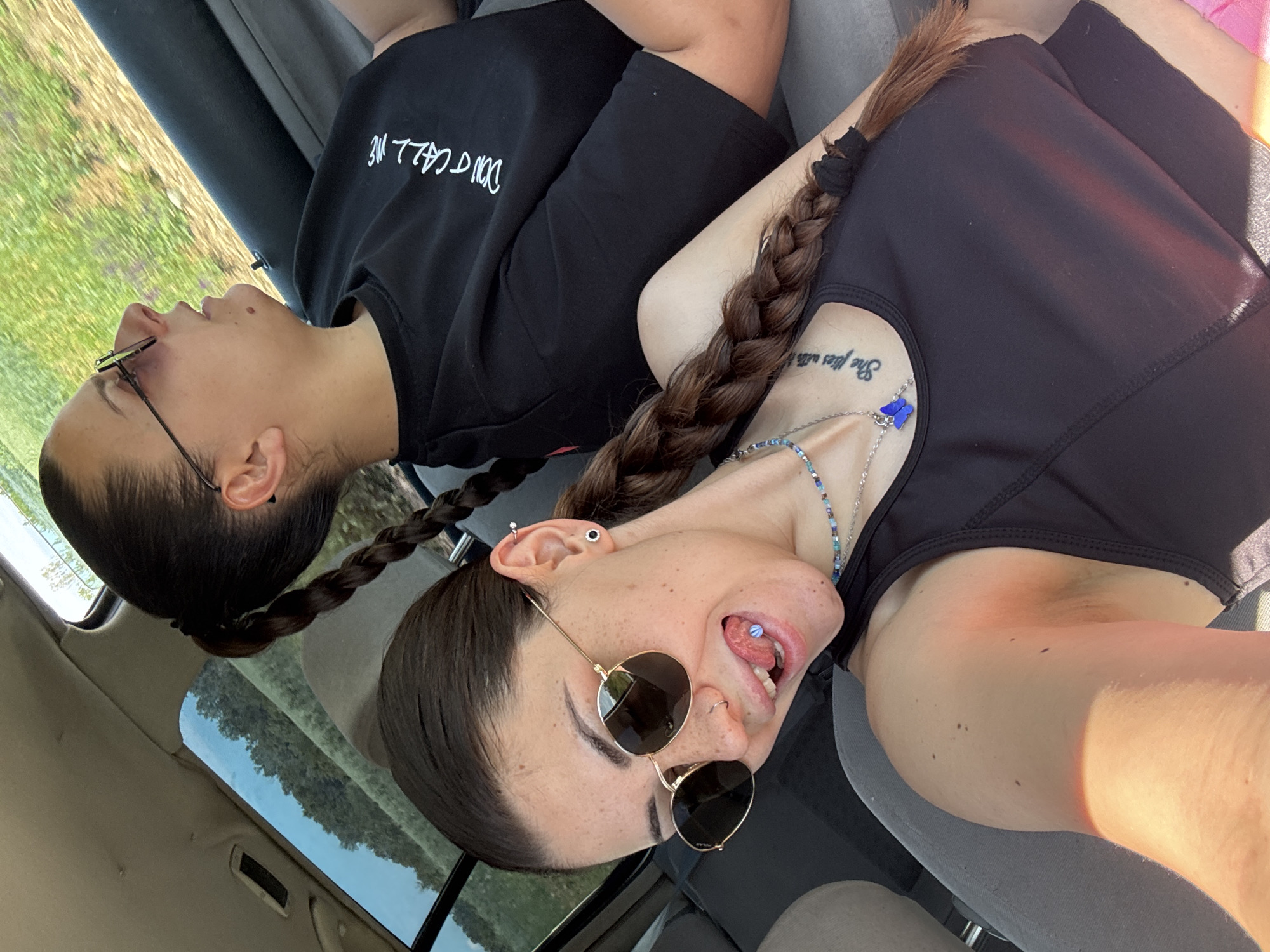 This media file captures two young women inside a car, likely reclining in the front seats, under bright daylight conditions in Belopoptsi, Bulgaria. The image is oriented sideways, with the subjects appearing to lie on their backs.
Both women have long, dark hair styled in thick braids. The woman on the left wears eyeglasses and a black long-sleeve top or hoodie with white, inverted text that reads "DON'T CALL ME". Her face is in profile, looking upwards with a serene expression.
The woman on the right wears round sunglasses and a black tank top. She has multiple ear piercings and a blue stud visible in her tongue piercing as she opens her mouth, suggesting a playful or expressive mood. Around her neck, she wears a multi-colored beaded necklace with a prominent blue butterfly charm. A tattoo on her upper chest/shoulder, written in a script font, is partially visible and appears to read "She flies with...".
The vehicle's interior features grey seats and a beige headliner. Through the windows and possibly a sunroof, a clear blue sky and verdant green trees are visible, indicating a sunny outdoor setting. Strong sunlight casts clear shadows on the subjects, highlighting the bright conditions.