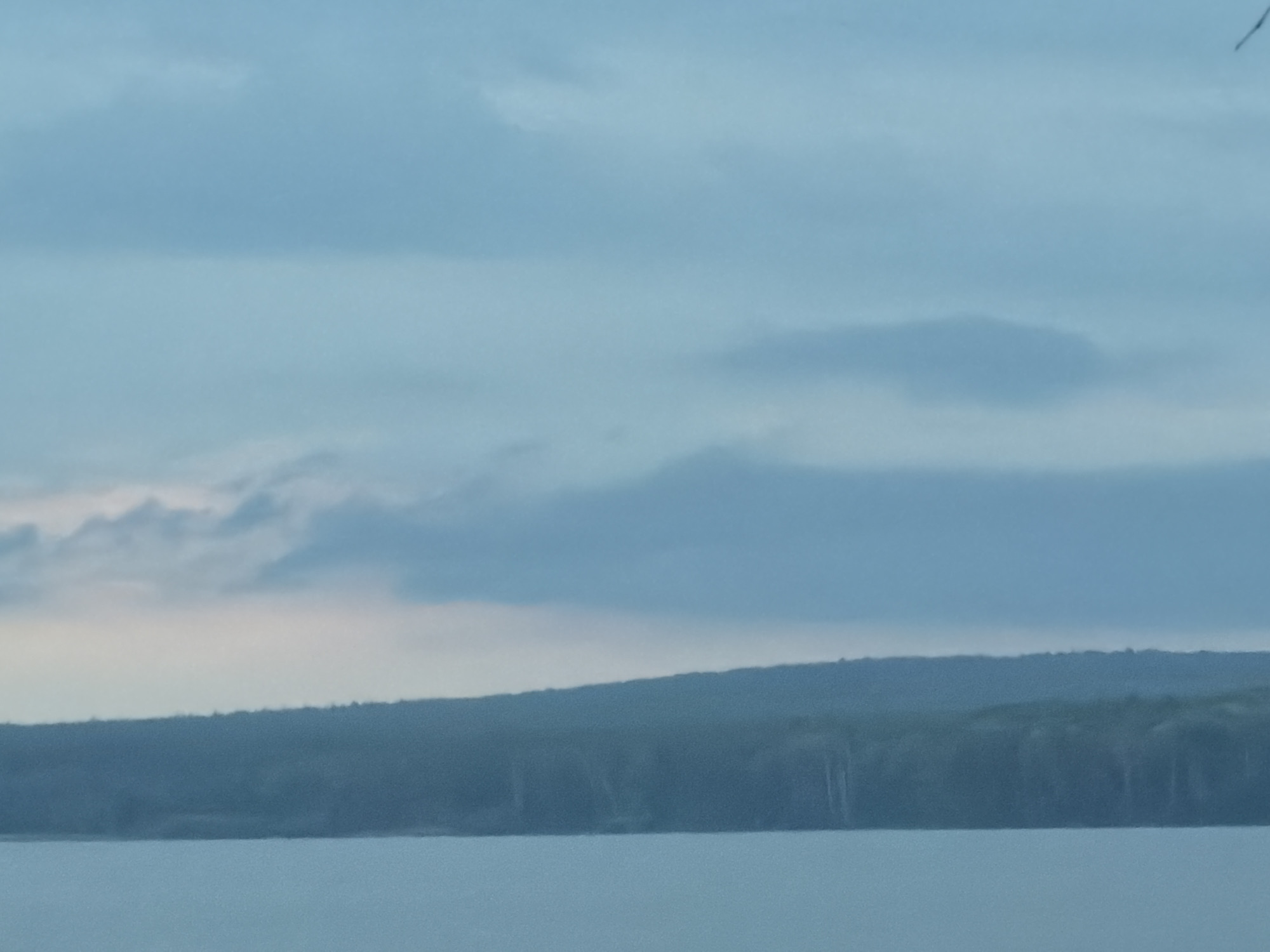 The image depicts a body of water in the foreground, likely a lake or wide river, with a line of trees and a low, rolling hill on the horizon. Above, the sky is filled with clouds. The lighting is subdued, suggesting either early morning or late evening.
The exact location is not determinable from the image alone; the reference to Chaikovsky, Russia is not evident within the frame.