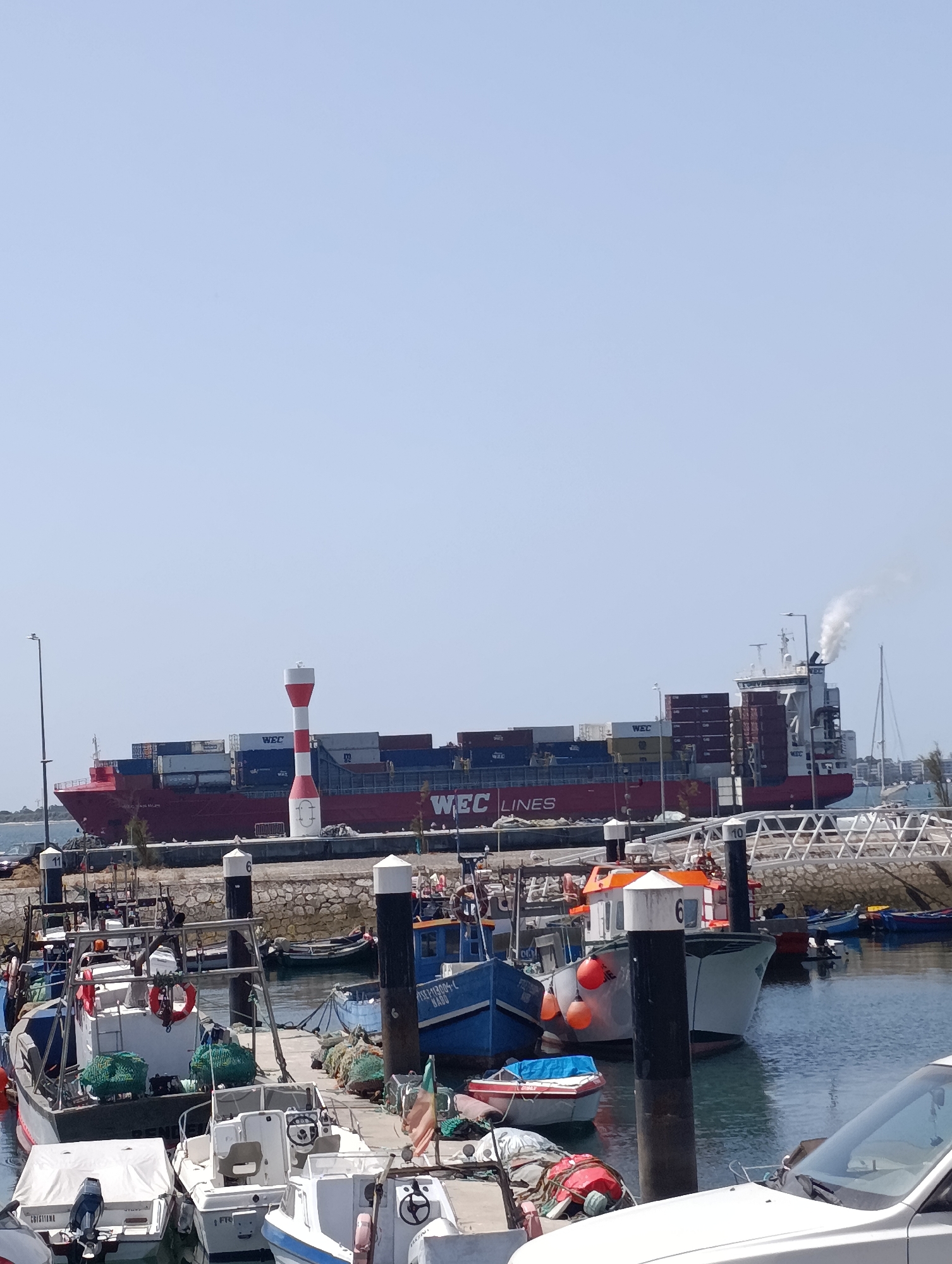 This image depicts a harbor scene in Setúbal, Portugal, with a large container ship, the WEC Lines, prominently featured in the background. The ship is red and laden with containers, some of which are dark blue. Smoke billows from the ship's funnel, suggesting it is operational. In the foreground, several smaller boats are docked. One boat is white with a blue hull, another is white and covered with a tarp, and a third appears to be a small, white motorboat. A red and white striped lighthouse or marker stands near the container ship. The water is calm and reflects the blue sky. The dock area is visible, with bollards and some netting. A white car is partially visible in the lower right corner of the image. The overall scene suggests a working port on a clear, sunny day.