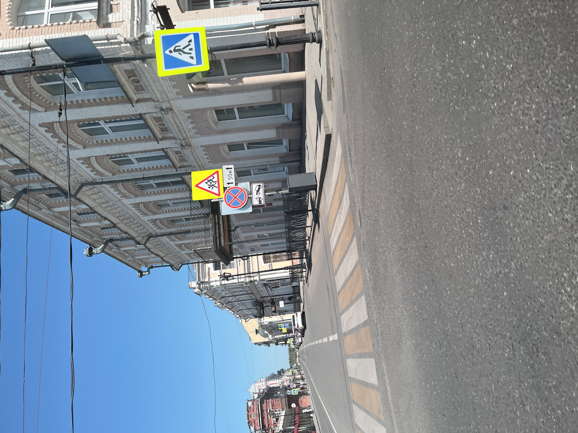 The image depicts a street scene in Astrakhan, Russia, on a clear, sunny day. The perspective is from a vehicle on the road, looking down a street lined with multi-story buildings. A pedestrian crossing is prominently in the foreground.
Key elements in the scene include:
**Buildings:** The buildings are a mix of architectural styles, with some featuring classical elements like arched windows and decorative facades. One building, possibly older, on the left has red brickwork and a dome-like structure. Another, on the right, is lighter in color with intricate white detailing around the windows. Balconies with railings are visible on some of the buildings.
**Road and Street Furniture:** The road surface appears to be asphalt. Road markings include a pedestrian crossing with white and yellow stripes. Several traffic signs are visible on poles. These include:
* A yellow triangular sign with two children running.
* A circular red and blue sign indicating no parking or stopping.
* A rectangular sign with an upward arrow and "50МТ" (likely indicating 50 meters).
* A blue square sign with a white pedestrian symbol.
A black trash receptacle is positioned near the sidewalk. Streetlights are also visible.
**Vehicles:** Several cars are present on the road. A white car is particularly noticeable in the middle distance.
**Environment:** The sky is a clear, bright blue. Overhead wires are strung across the street, supported by poles. The overall impression is of a well-maintained urban environment on a pleasant day.
The scene is static, with no clear actions or interactions depicted, other than the implied movement of traffic on the road. The image captures a typical urban streetscape with elements of daily life, such as pedestrian crossings and traffic regulation signs.