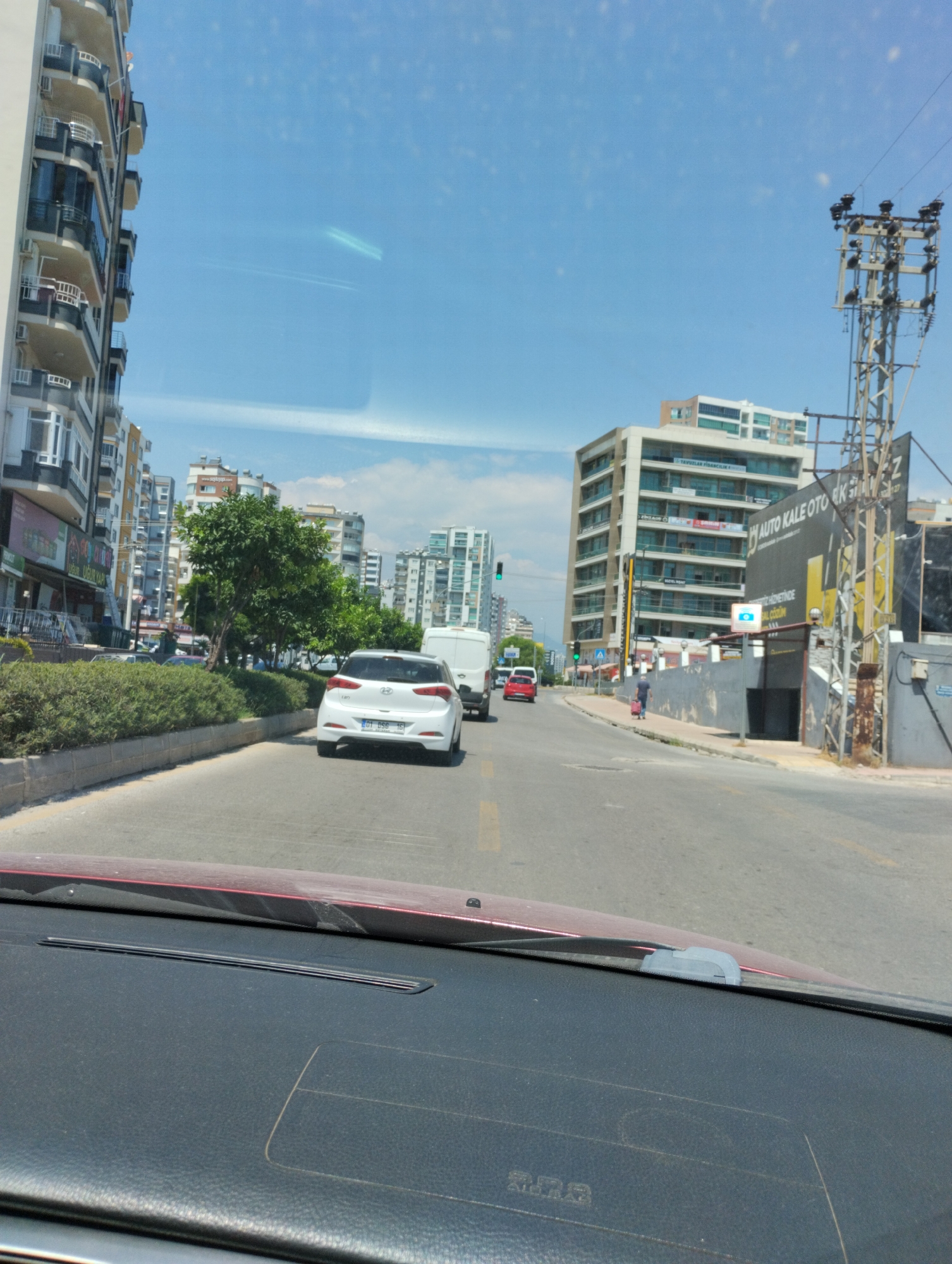 The image depicts a street scene in Mezitli, Turkey, likely during daytime with clear skies. The perspective is from inside a vehicle, looking forward. A white hatchback is in the foreground, driving on a road with painted lane markers. To the left, a tall, multi-story apartment building with balconies is visible. To the right, there's a utility pole and a larger, modern building with signs, one of which reads "AUTO KALE OTO EKSPERTIZE". Further down the street, other buildings and trees line the road. A few other vehicles, including a white van and a red car, are also present on the road. A person is walking on the sidewalk with a wheeled suitcase. The overall environment suggests an urban setting with commercial and residential buildings.