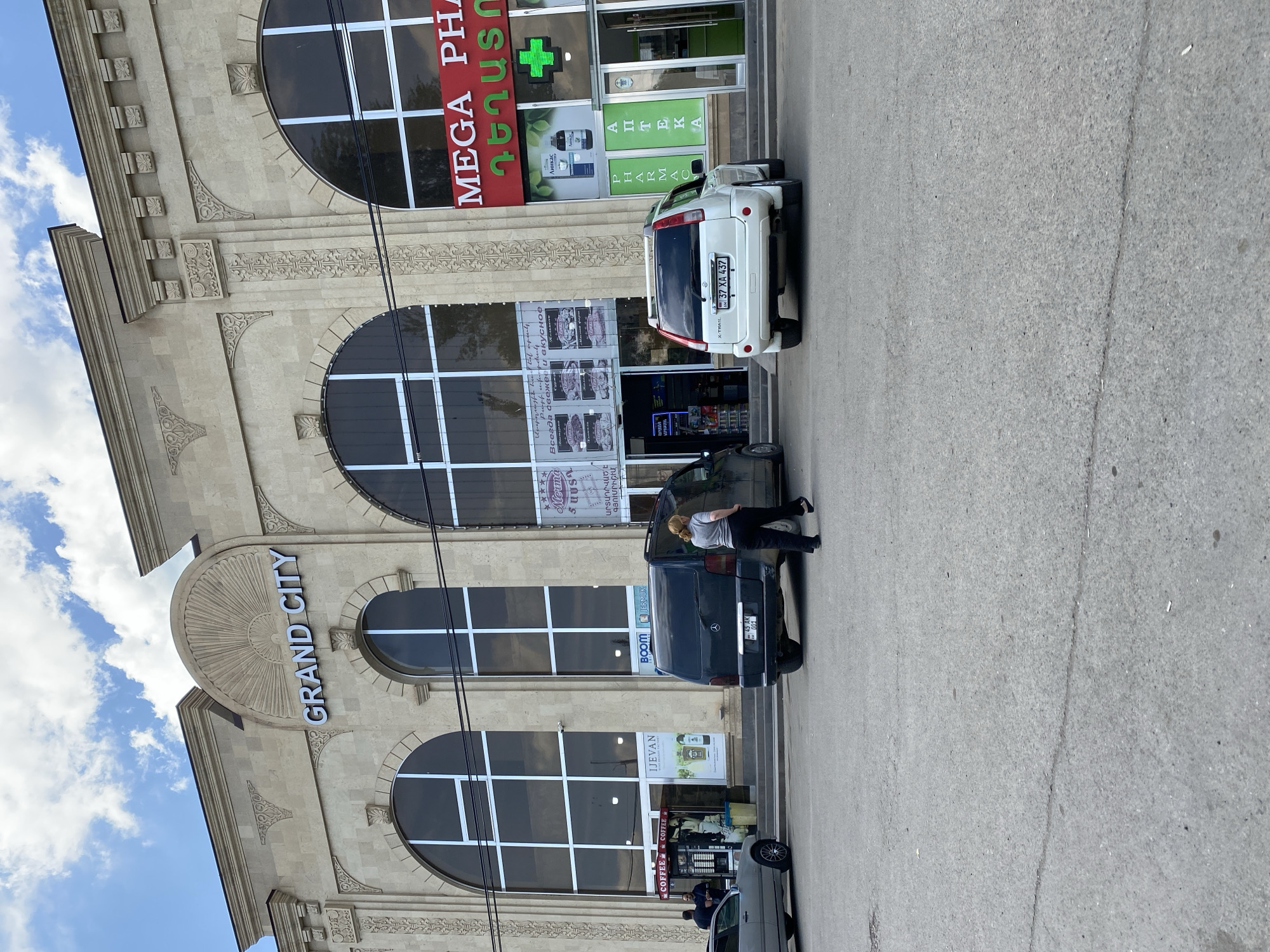 The image shows the exterior of a building with the prominent signage "GRAND CITY." The architecture features large arched windows and decorative stonework. To the left of the entrance, there is a "COFFEE" stand. Further to the right, there is a pharmacy with red signage that reads "MEGA PHARMA" and a green cross symbol. Several vehicles are parked in front of the building, including a dark minivan and a white SUV. A woman is walking away from the camera in the foreground, near the white SUV. The scene appears to be outdoors on a paved area, under a partly cloudy sky, and the building is located in the city of Gyumri, Armenia.