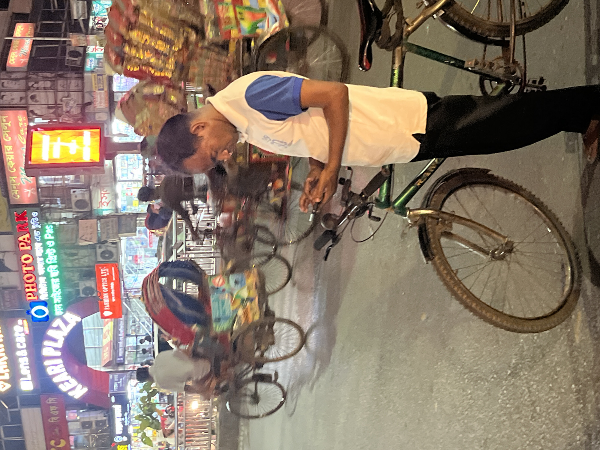The scene is a nighttime street view in Dhaka, Bangladesh. A man is standing next to a green bicycle, seemingly working on it. He is wearing a white shirt with blue sleeves and black pants. In the background, there is a cluttered urban environment with numerous shops and signs. Key signage includes "PHOTO PARK" and "KEKARI PLAZA" in prominent, illuminated displays. The area seems busy with other pedestrians and cyclists. The lighting is a mix of artificial light sources, creating a vibrant atmosphere.