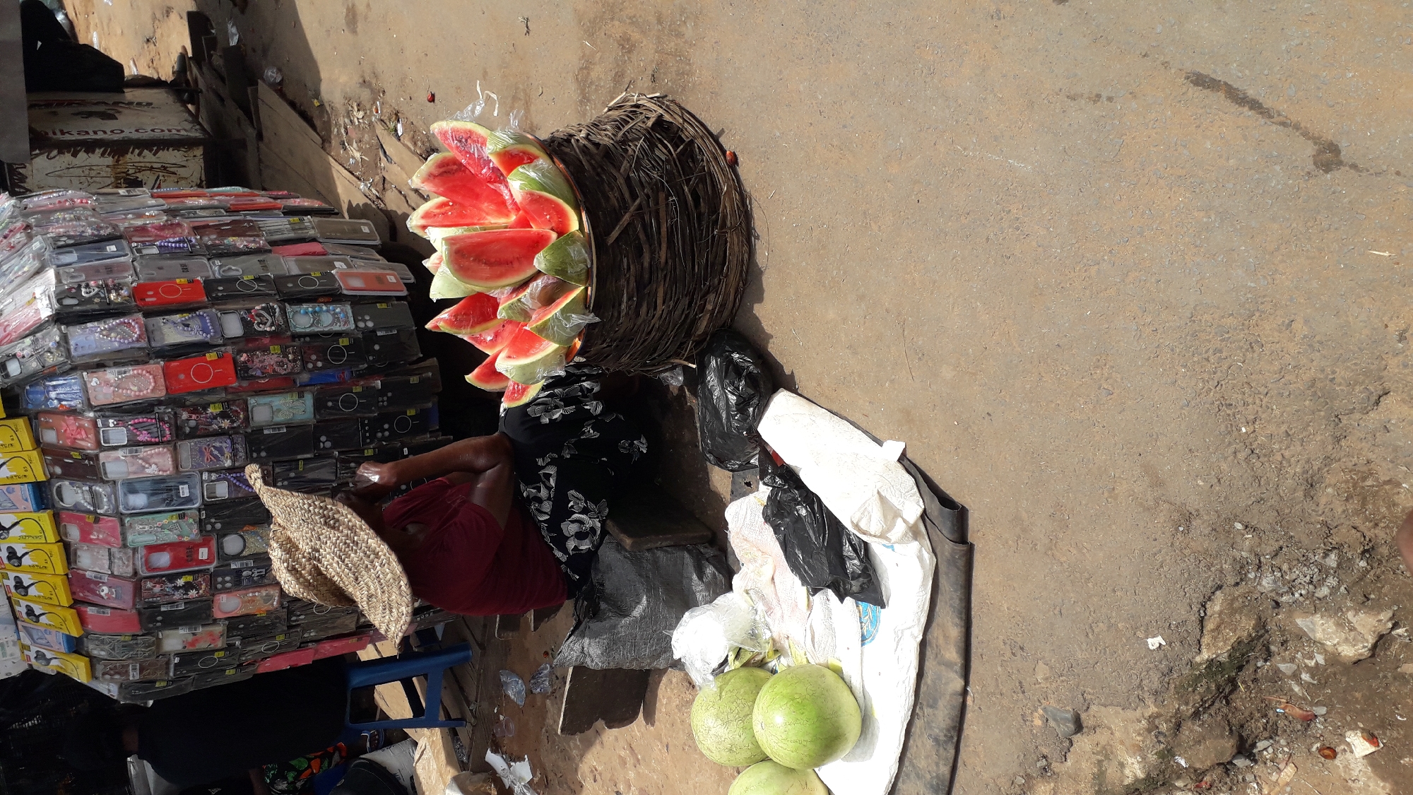 In Nkpor, Nigeria, a woman wearing a straw hat and a patterned black and white skirt sits on a blue plastic stool next to a stack of packaged phone cases. A large woven basket filled with slices of watermelon sits beside her. The watermelon slices are wrapped in clear plastic. Next to the basket are black plastic bags and a white cloth. Three whole watermelons sit on the cloth. The scene is on a concrete surface in an outdoor location.