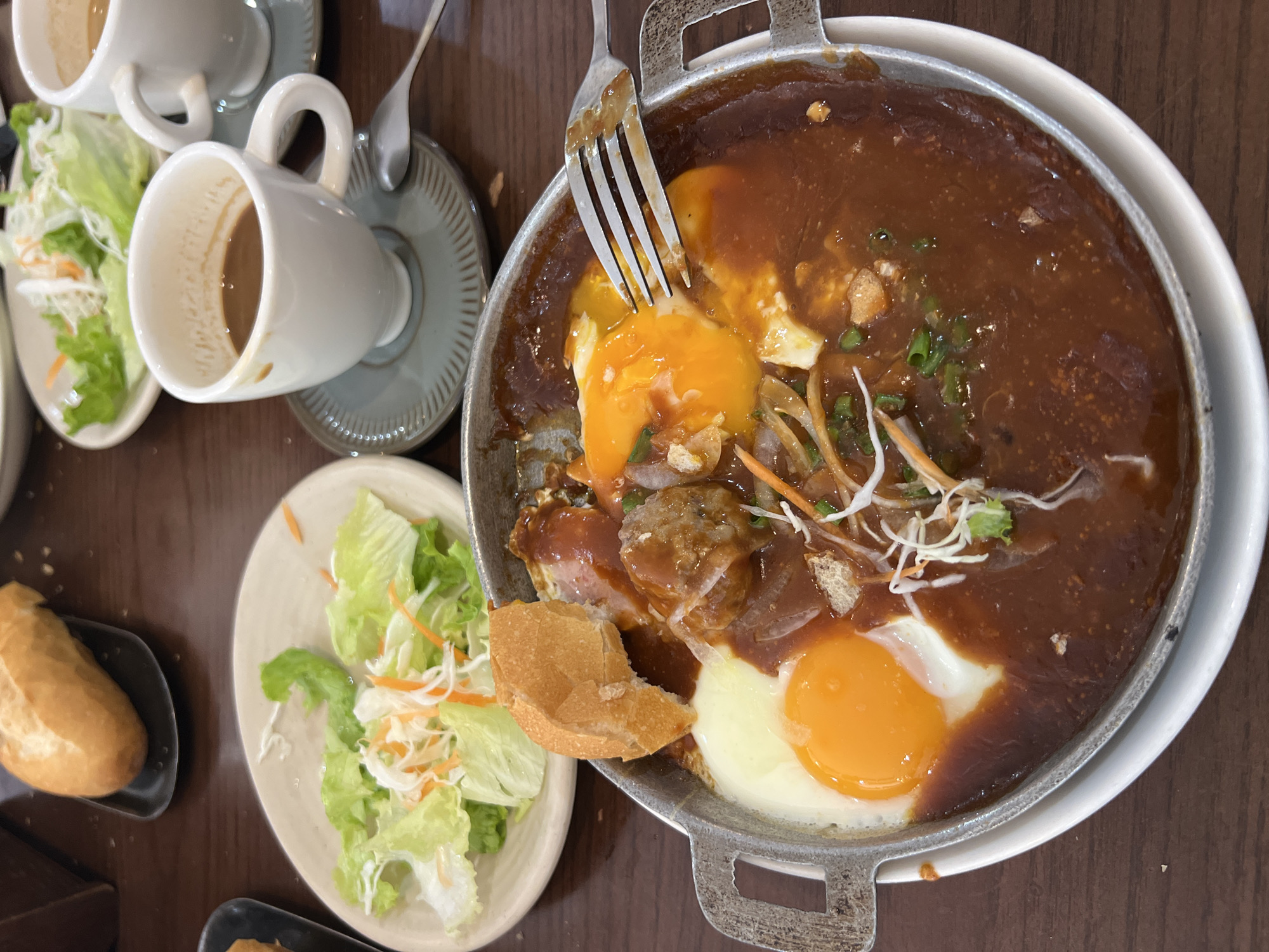 In an overhead shot, a meal is presented on a dark wood table. The focal point is a sizzling skillet containing two fried eggs, a meatball, and a piece of bread, all bathed in a rich, dark sauce. The skillet is placed on a white plate. To the left of the skillet is a small white plate with a salad of lettuce and shredded carrots. In the background, two white coffee cups sit on dark gray saucers, with a spoon resting on one of them. The cups appear to be partially filled with coffee. Another piece of bread sits on a black dish behind the salad. The scene suggests a casual dining experience.