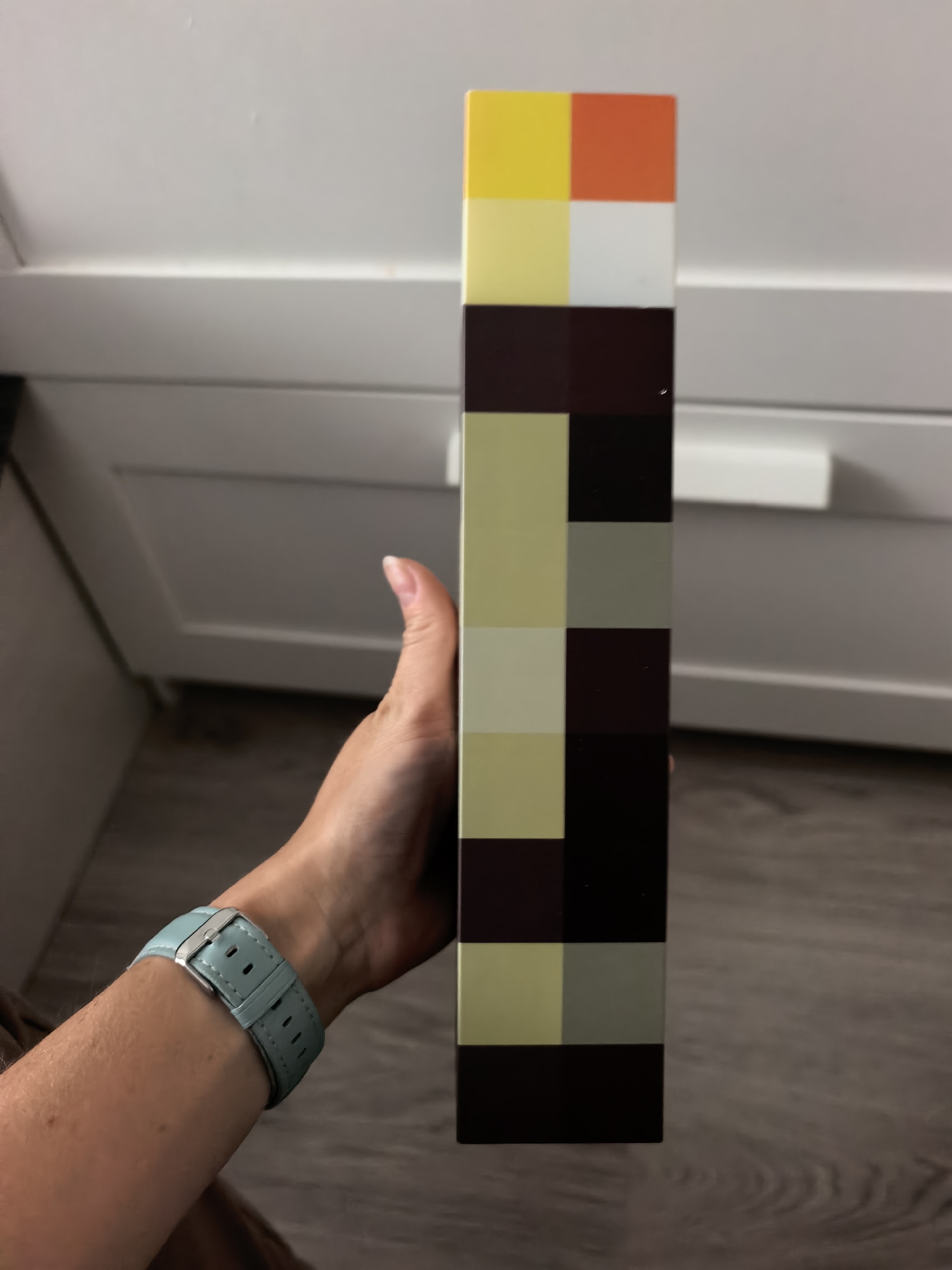 The image shows a person's hand holding a rectangular object with a checkered pattern of colored squares. The object is predominantly made up of squares in shades of yellow, white, orange, brown, and gray. The person is wearing a light blue watch on their wrist. The background appears to be an indoor setting with white furniture, possibly cabinets or drawers. The floor is a light-colored wood-grain pattern. The image does not contain any information that would allow for its location in Sterlitamak, Russia.