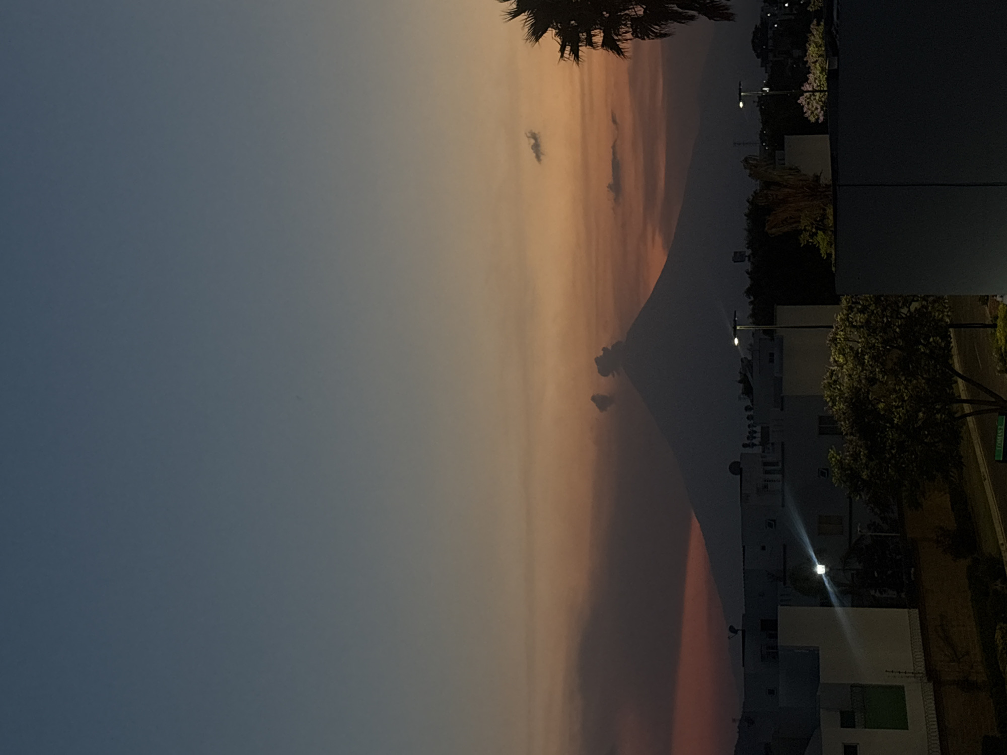 A vertical photograph displays a nighttime scene in San Bernardino Tlaxcalancingo, Mexico, with the sky dominating the left side. The sky transitions from a dark blue on the left to orange and pink hues closer to the right, suggesting either dusk or dawn. There are a few wispy clouds scattered across the sky.
On the right side of the image are buildings and street lights, indicating an urban or suburban environment. Some trees are visible, and a portion of a palm tree is present at the top. The illumination suggests it is not completely dark, implying the presence of artificial light sources in the area. The overall setting is a residential or mixed-use neighborhood at twilight or dawn.