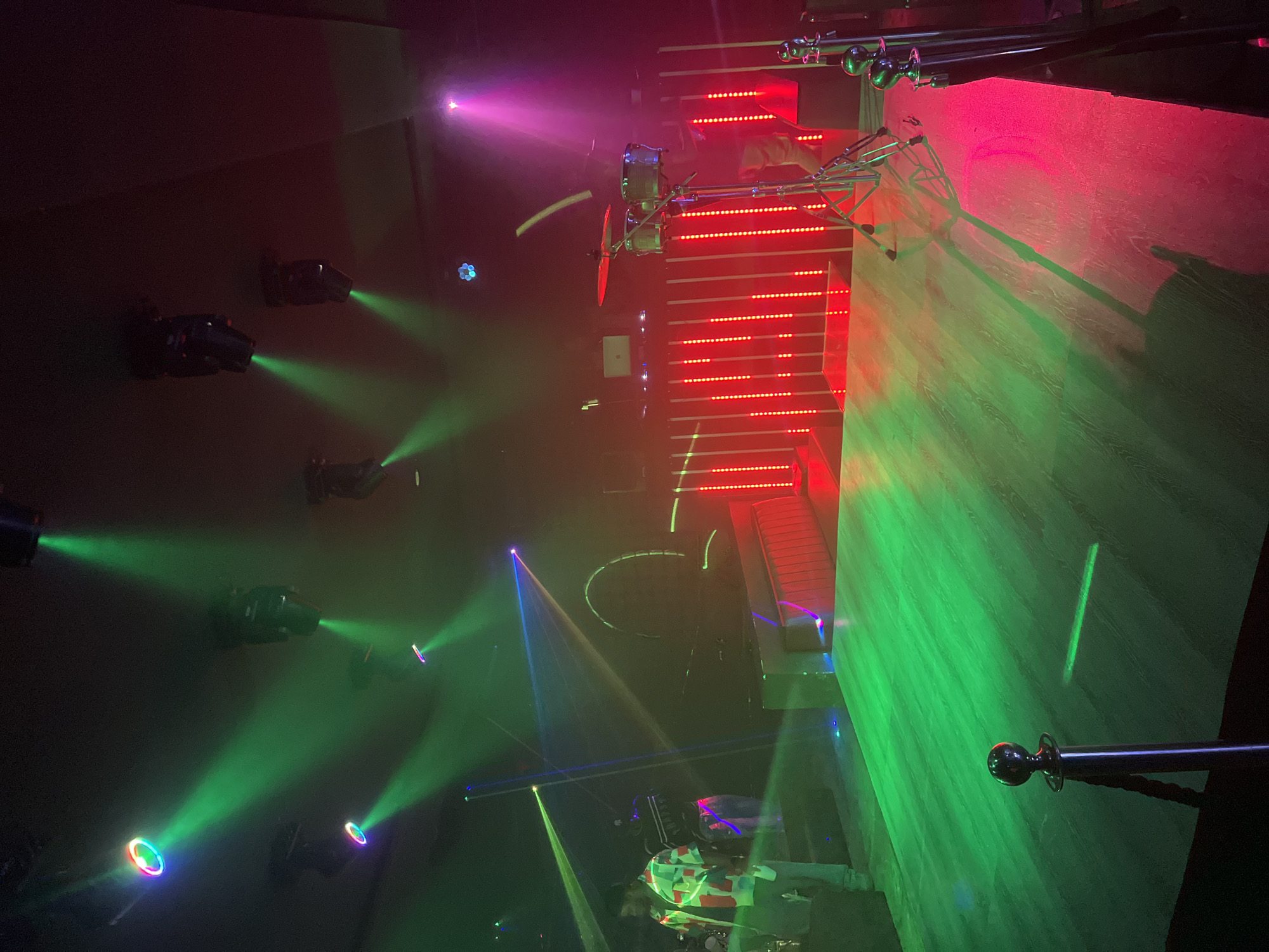 The image depicts an interior scene, likely a nightclub or entertainment venue, in Jalingo, Nigeria. The location is illuminated by a dynamic light display featuring a combination of red, green, pink, yellow, and blue beams and spotlights.
Several light fixtures are visible, casting beams across the room. Some fixtures are mounted overhead, while others are positioned on the floor.
A structural element, possibly scaffolding or a stage, is present and is highlighted by red lights. There is also a silver railing.
At least one individual is visible, located towards the bottom of the image, though details about the person are difficult to ascertain due to the lighting conditions. A set of drums are visible as well.