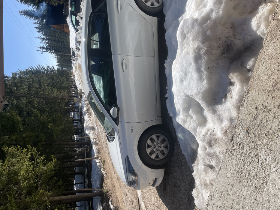 A white four-door hatchback vehicle, consistent with a Hyundai i20 model, is parked on the dirt and gravel shoulder of a snow-lined road in Palež, Montenegro. The driver's side front door of the vehicle is slightly ajar. A significant accumulation of dirty, melting snow rests on the ground immediately adjacent to the car and to the right of the foreground. The road, partially obscured by snowdrifts, extends into the distance through a dense coniferous forest. Patches of snow are visible on the forest floor. In the background on the right, a dark-roofed building, possibly a cabin or lodge, is visible, emitting smoke or steam from its chimney. Overhead utility lines are also present. The sky is clear and blue, indicating bright daylight. No individuals are visible in the scene.