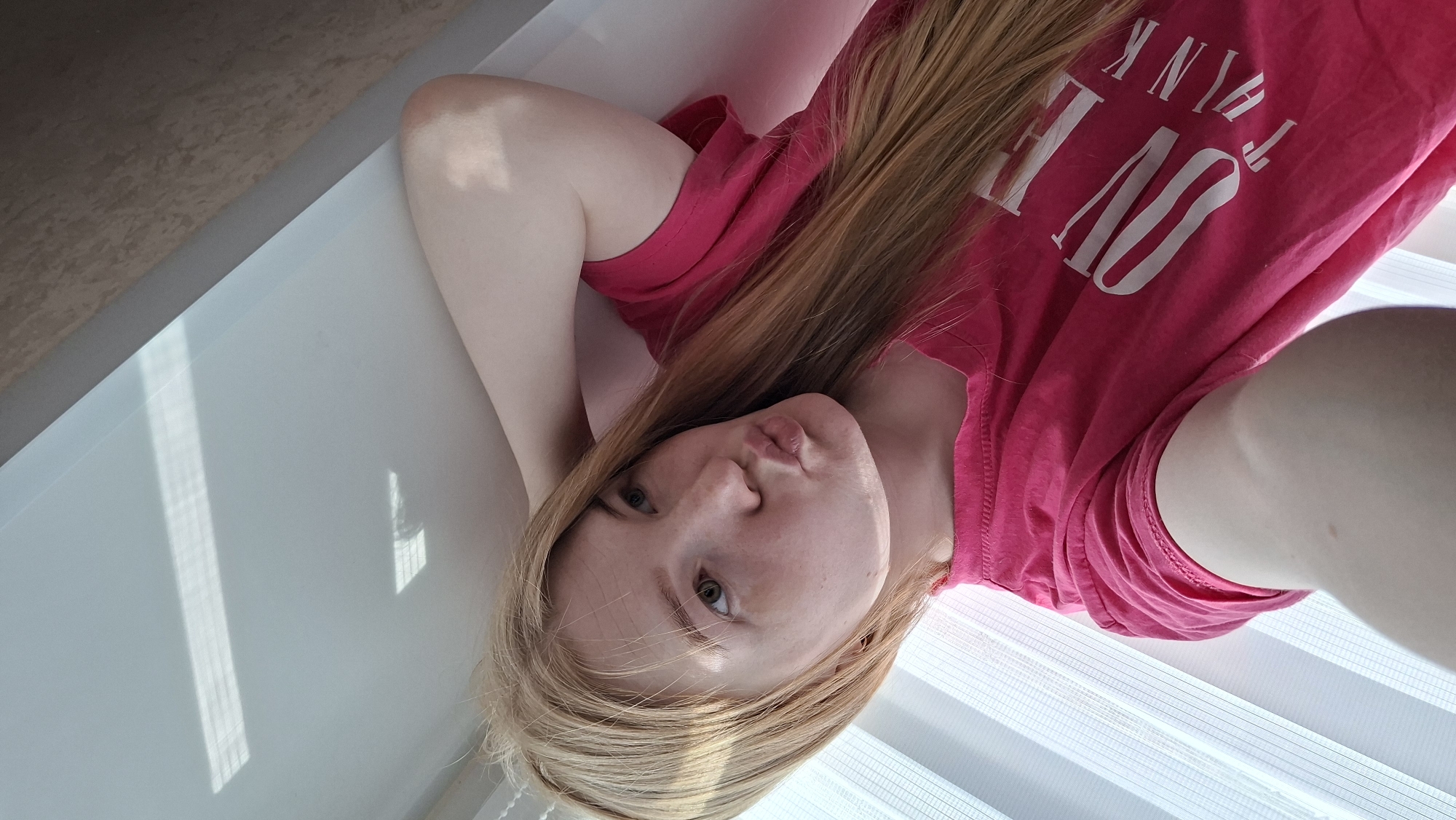 A young woman with long blonde hair is lying down in a bathtub. She is wearing a pink t-shirt with white lettering. The setting is a bathroom, with a window with blinds. The location is Chaikovsky, Russia.
